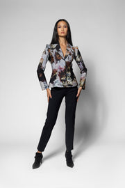 A woman modeling a long-sleeve jacket with a complex multicolor floral and abstract print, paired with black trousers and black ankle boots, against a light gray studio background.