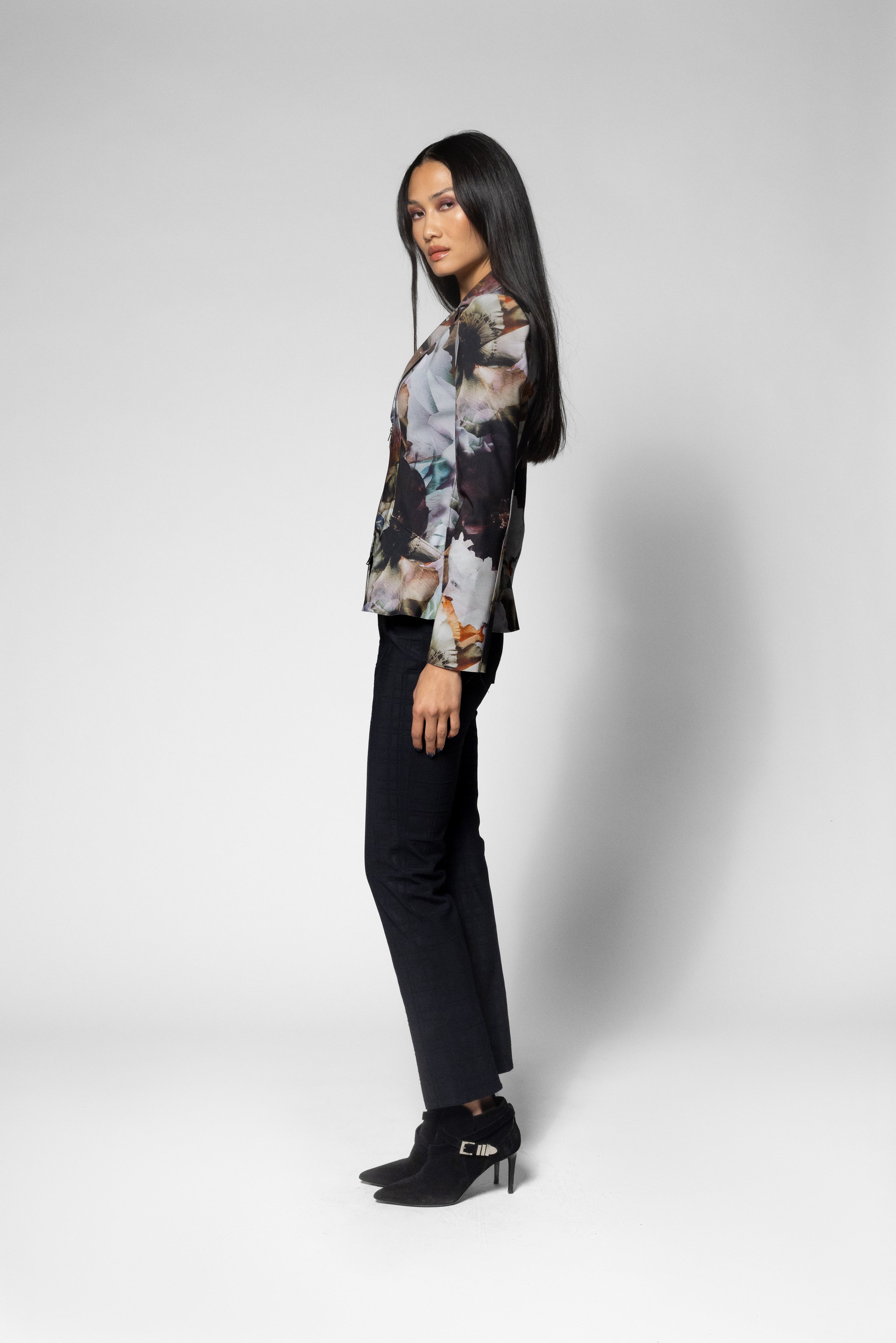 A woman stands in profile wearing a long-sleeve abstract print jacket with black pants and black ankle boots against a light gray studio background.