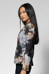 A woman with long black hair is shown in profile wearing a long-sleeve blazer with an abstract floral print in muted tones of gray, brown, beige, and black, paired with black pants, against a light gray studio background.