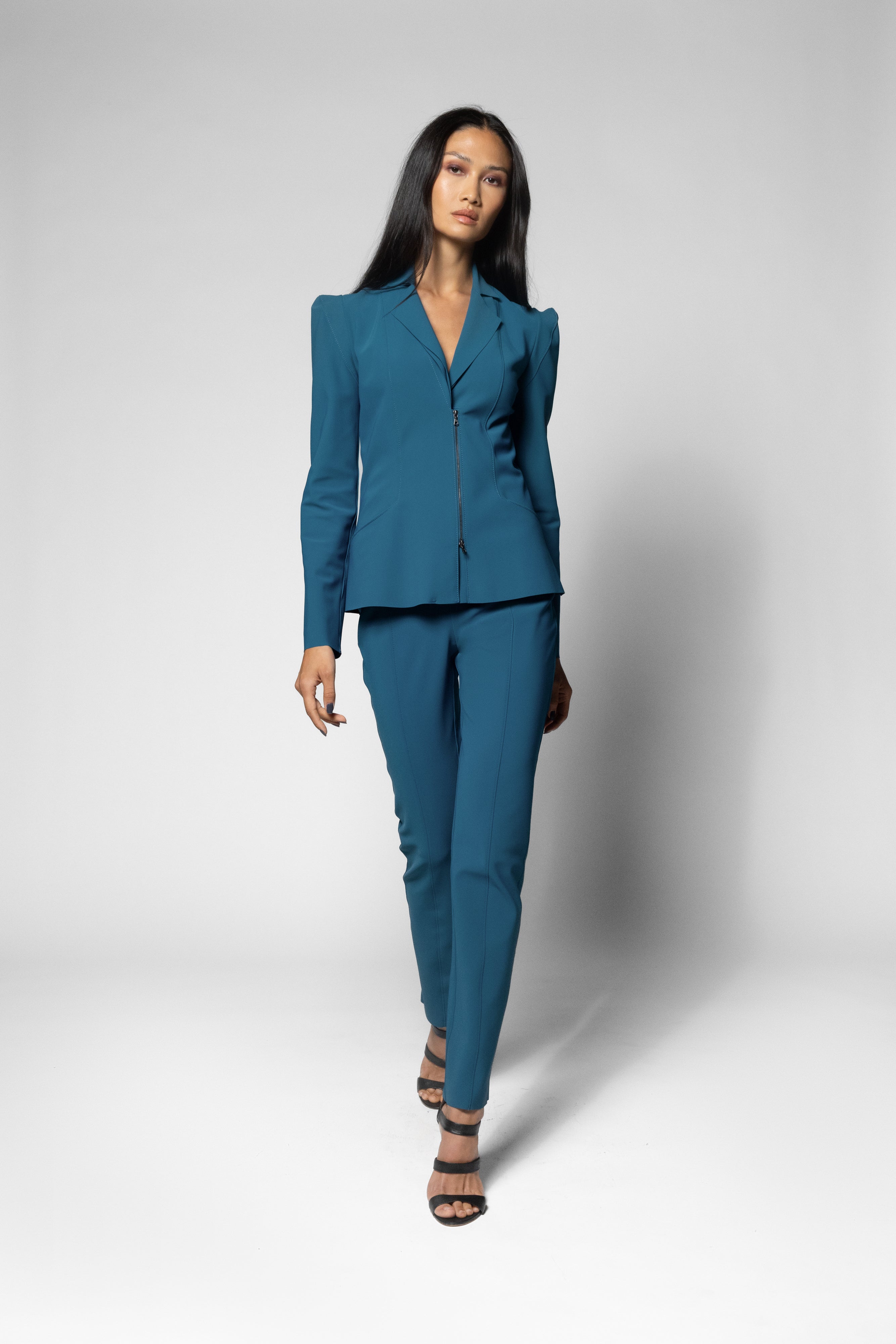 A woman modeling a teal blue tailored pantsuit with structured shoulders and a wrap-style front, paired with dark heeled sandals, against a light gray studio background.