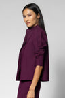A woman modeling a deep plum purple high-neck jacket with gathered elbow-length sleeves, paired with matching trousers, against a light gray studio background.
