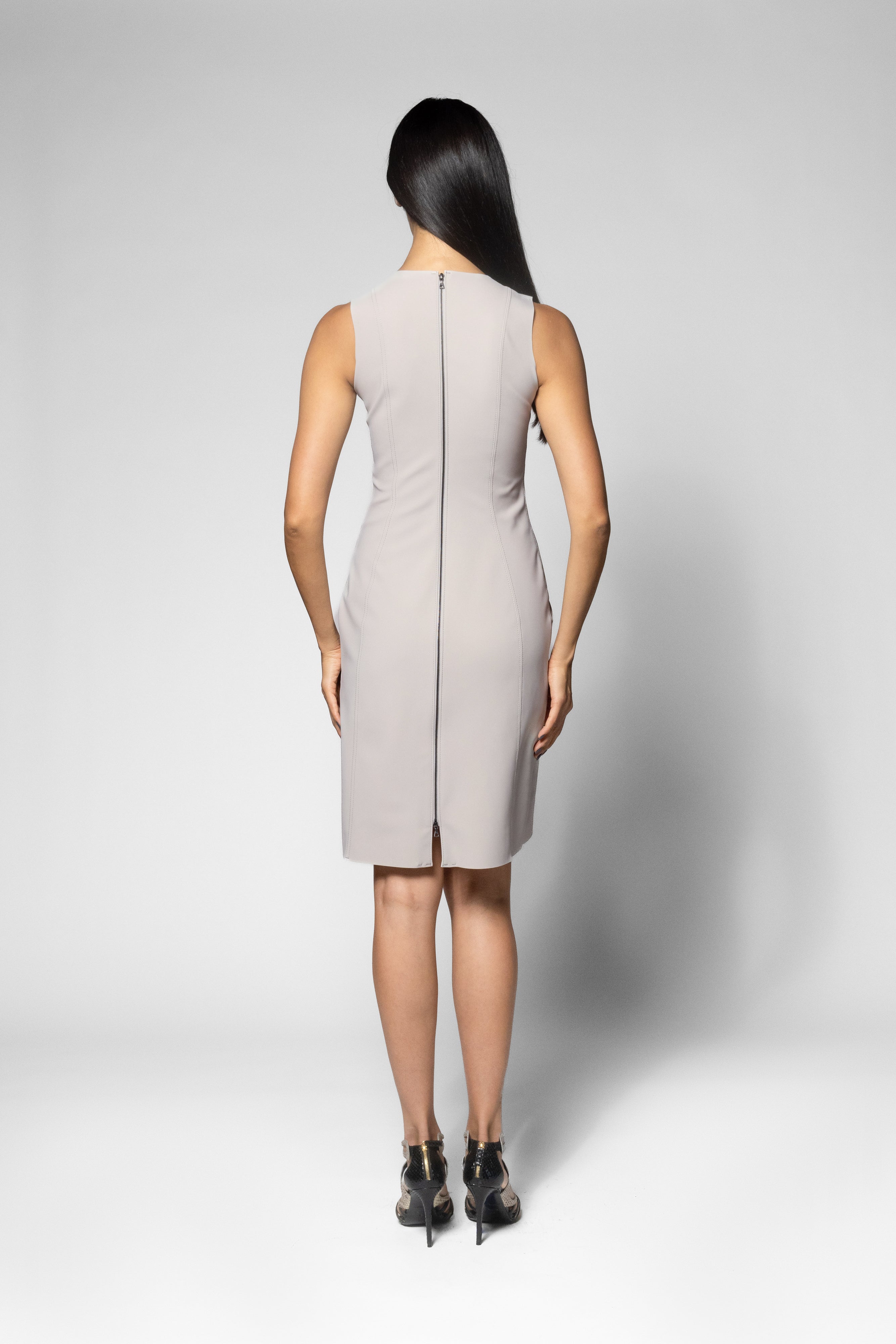 A woman viewed from the back wearing a sleeveless light beige knee-length dress with a central zipper, paired with black high heels, against a light gray studio background.