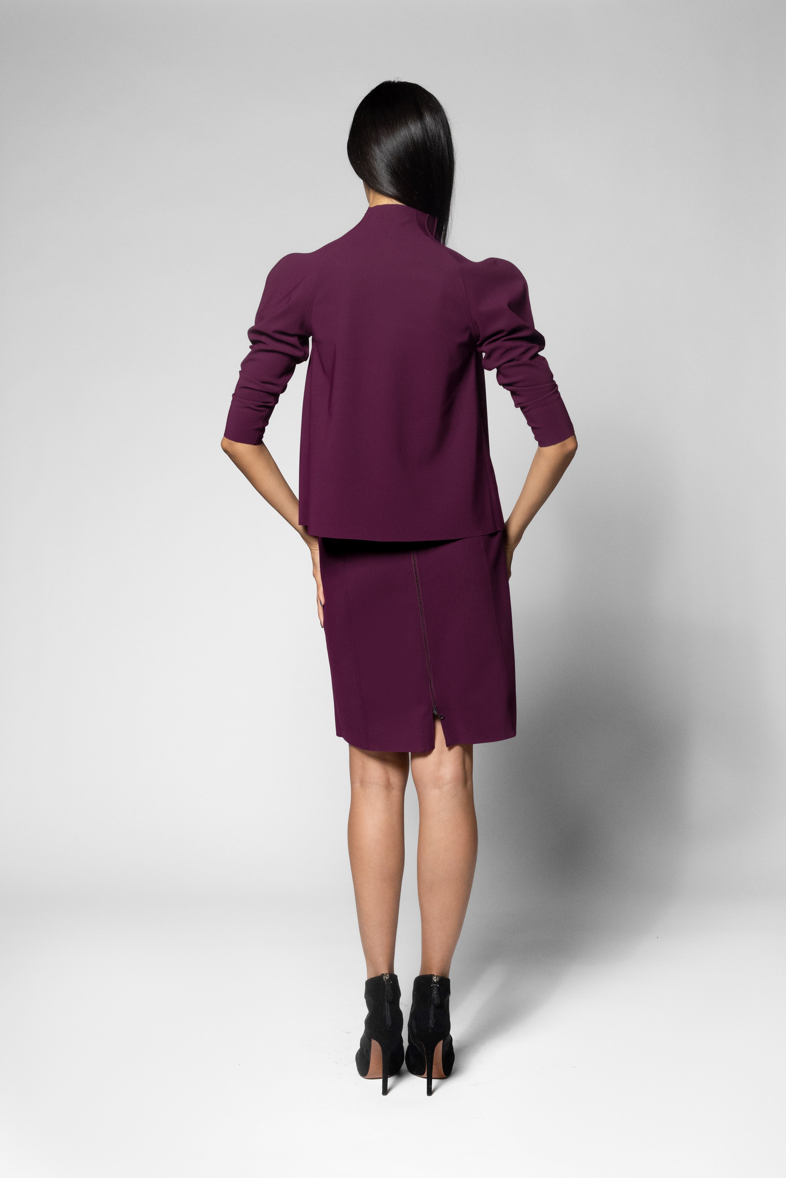 A model shown from the back wearing a matching deep purple blazer and skirt set, paired with black ankle boots, against a light gray studio background.