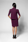 A model shown from the back wearing a matching deep purple blazer and skirt set, paired with black ankle boots, against a light gray studio background.