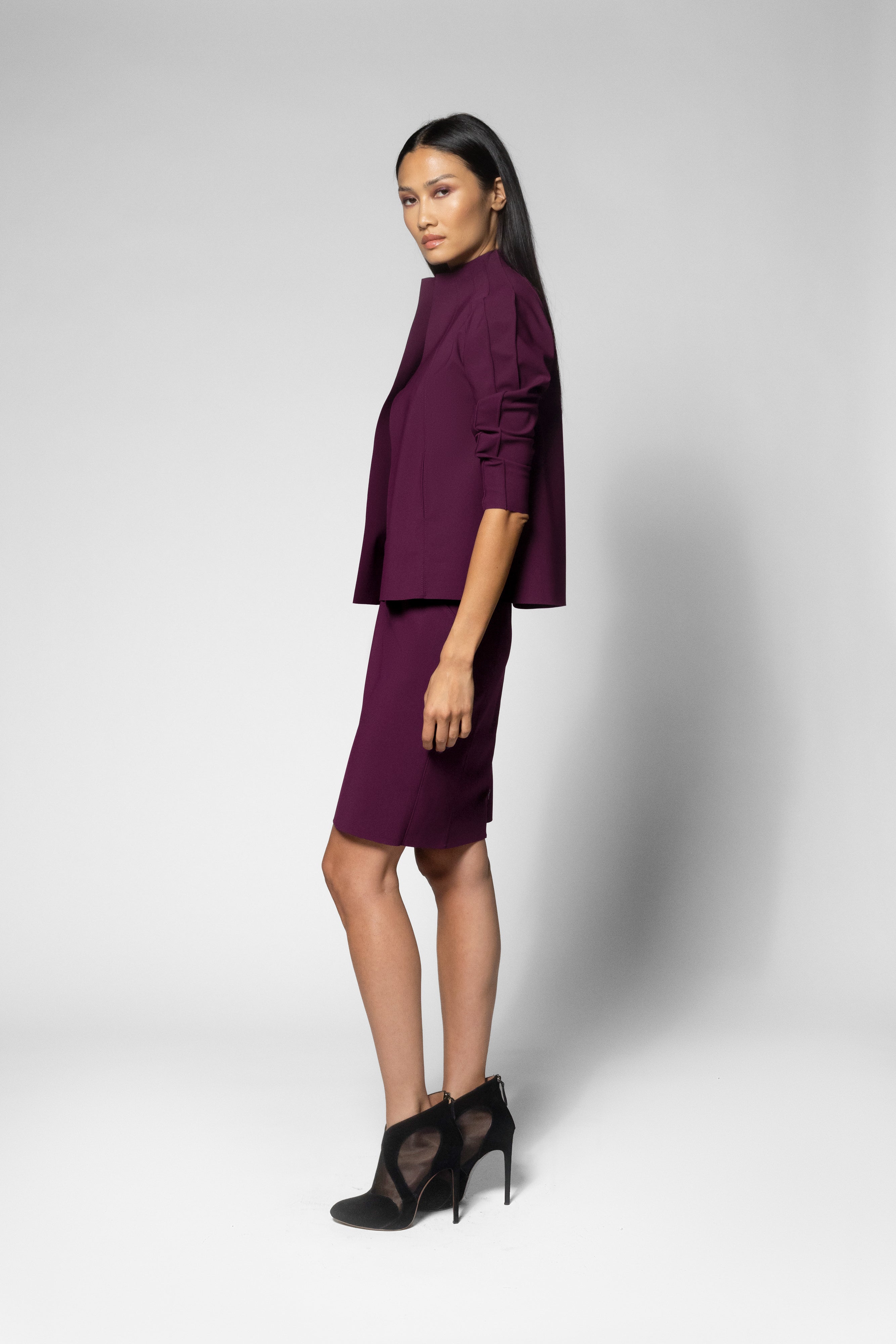A woman models a deep purple two-piece set consisting of a short-sleeve cropped jacket with ruffled detailing and a matching knee-length skirt, paired with black high-heeled shoes, against a light gray studio background.