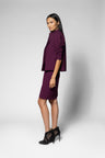 A woman models a deep purple two-piece set consisting of a short-sleeve cropped jacket with ruffled detailing and a matching knee-length skirt, paired with black high-heeled shoes, against a light gray studio background.