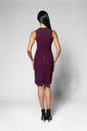 A woman viewed from the back wearing a sleeveless knee-length purple dress and black ankle boots, standing against a light gray studio background.