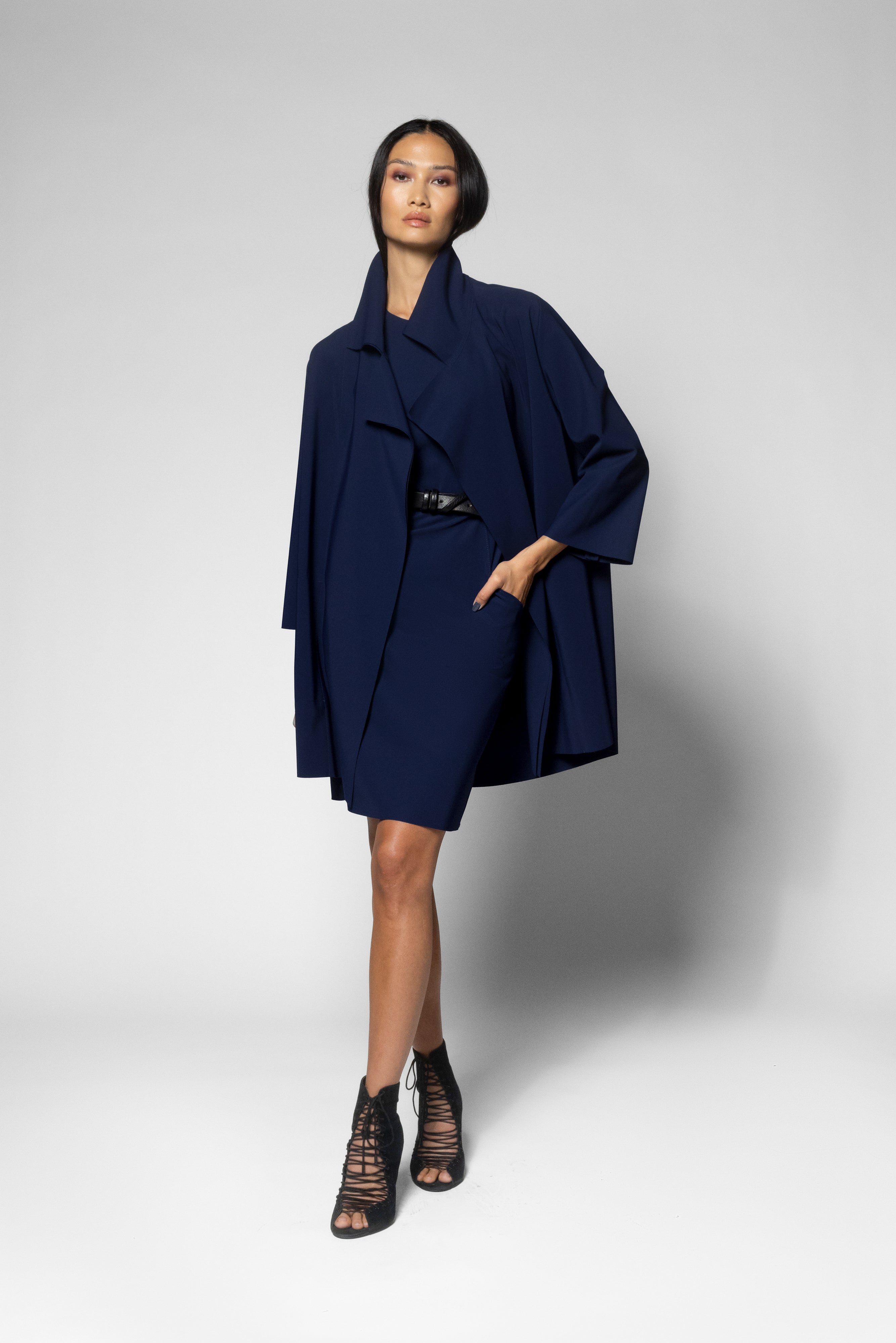 A model wearing a dark navy blue wrap-style coat with wide draped sleeves, a matching belt, and black lace-up ankle boots, standing against a light gray studio background.