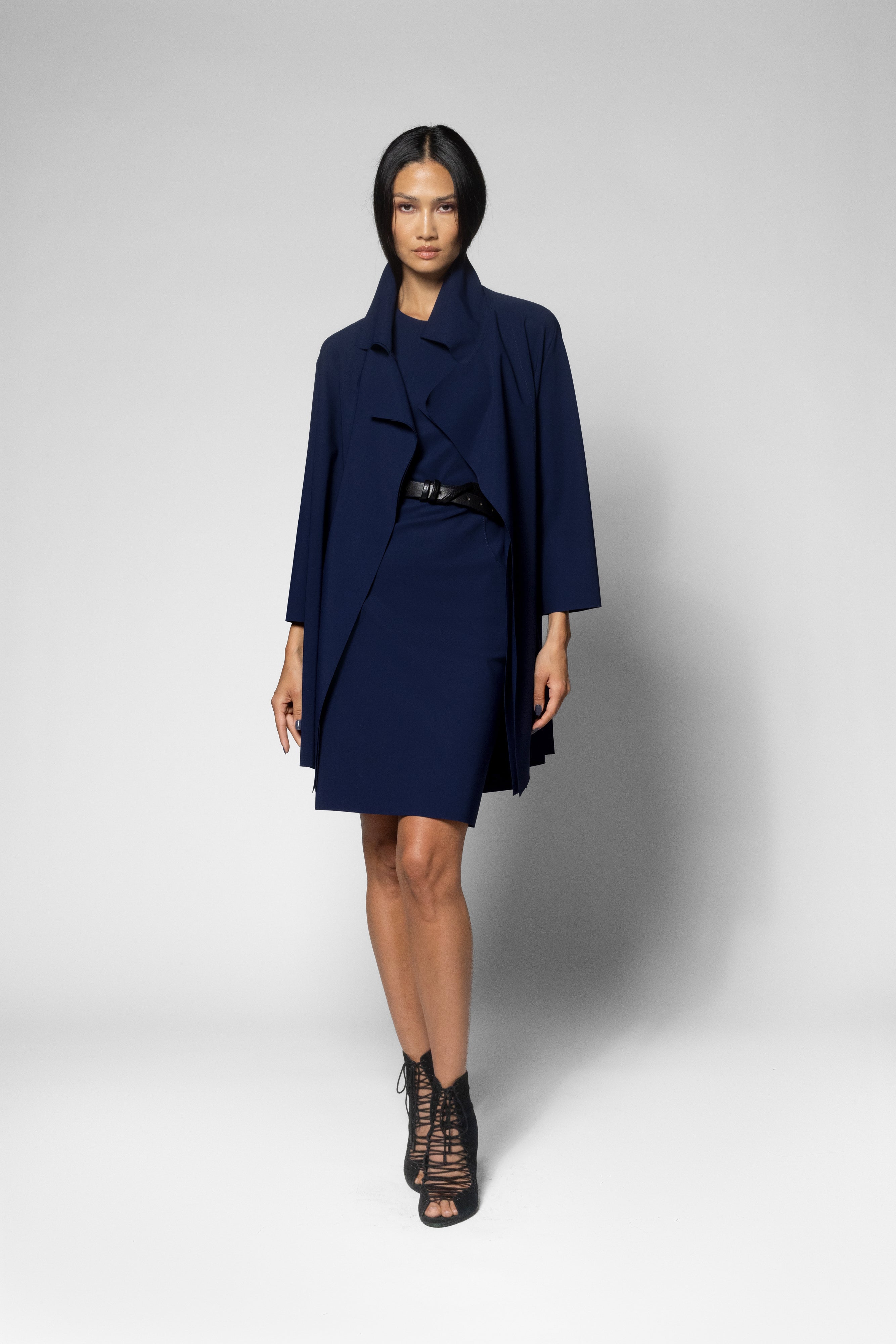 A model wearing a navy blue wrap-style coat over a matching navy dress, styled with a black belt and black lace-up ankle boots, against a light gray studio background.