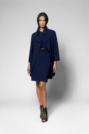A model wearing a navy blue wrap-style coat over a matching navy dress, styled with a black belt and black lace-up ankle boots, against a light gray studio background.