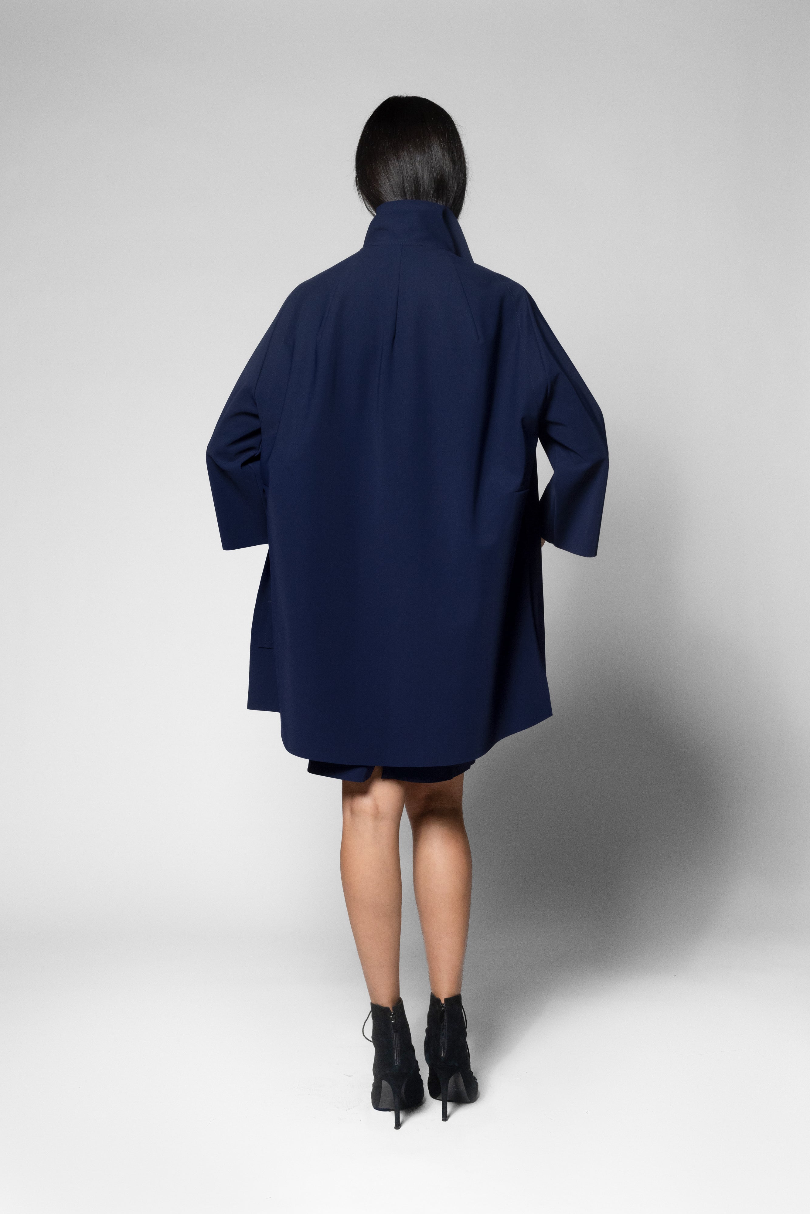 A model viewed from the back wearing an oversized navy blue coat dress with wide three-quarter sleeves and black ankle boots, photographed against a light gray studio background.