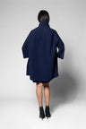 A model viewed from the back wearing an oversized navy blue coat dress with wide three-quarter sleeves and black ankle boots, photographed against a light gray studio background.