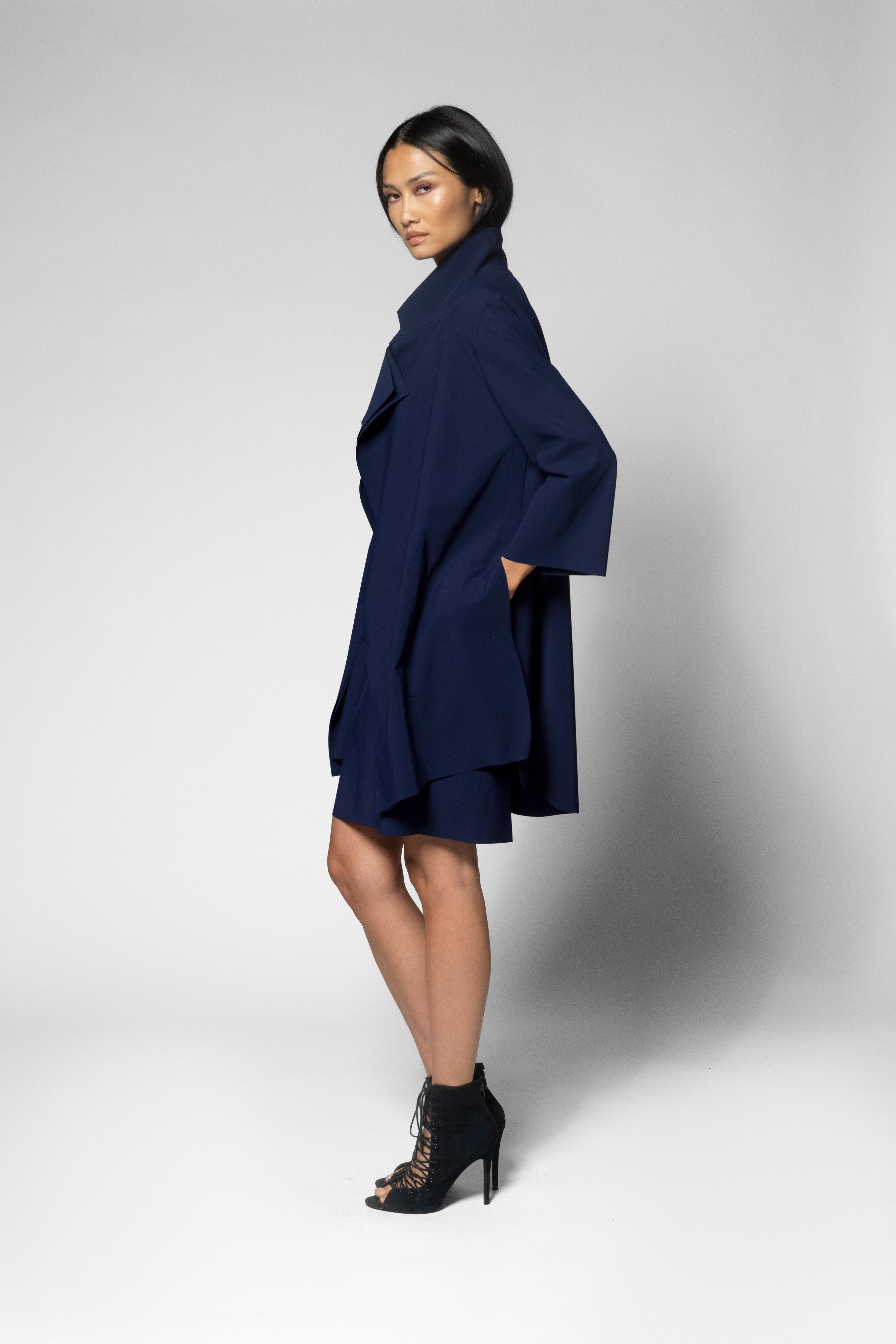 A model wearing a navy blue wrap-style coat dress with wide sleeves and a high collar, paired with black lace-up ankle boots, posed against a light gray studio background.