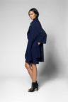 A model wearing a navy blue wrap-style coat dress with wide sleeves and a high collar, paired with black lace-up ankle boots, posed against a light gray studio background.
