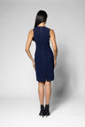 A woman viewed from the back wearing a sleeveless navy blue knee-length dress with a straight back and black ankle boots, standing against a light gray studio background.