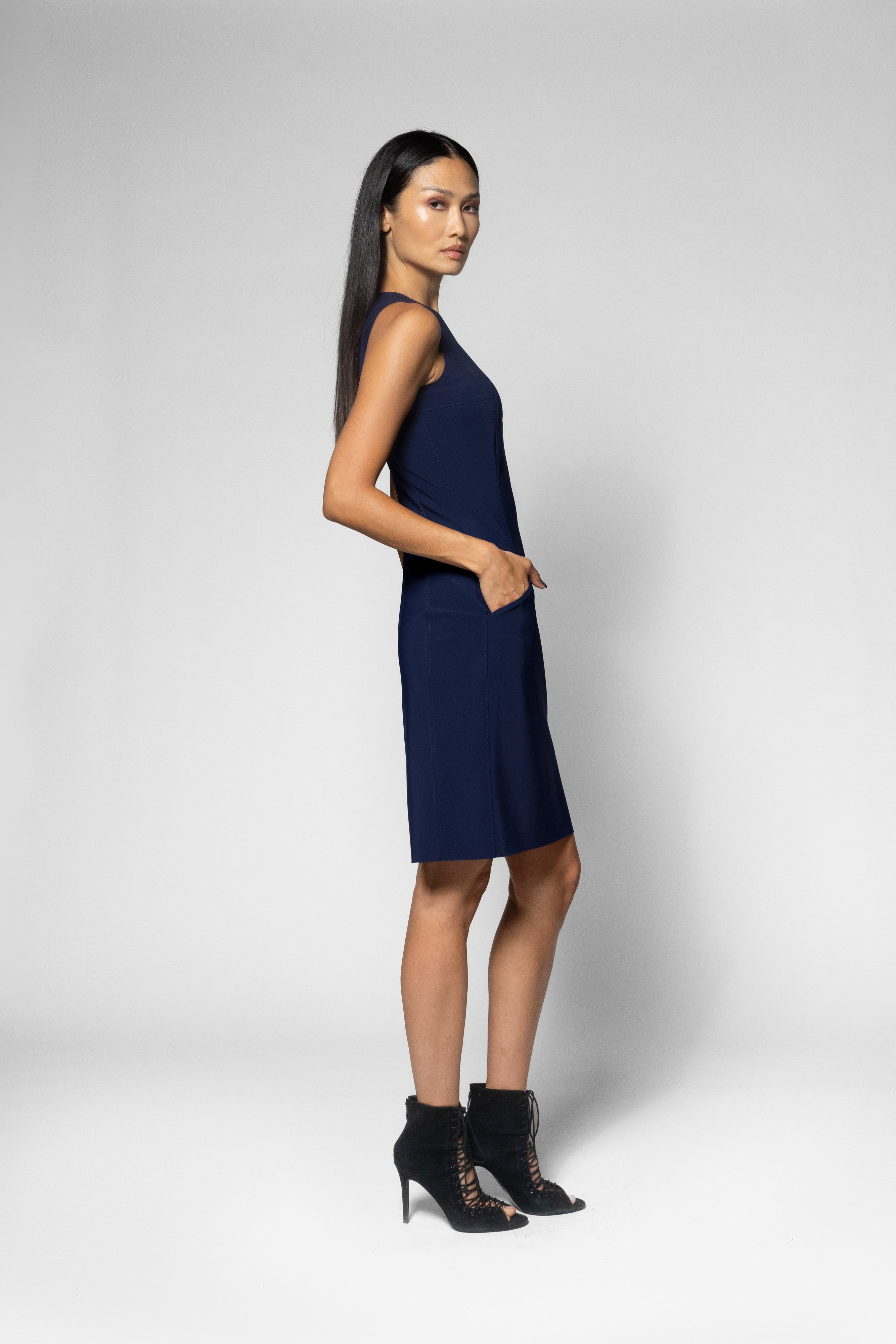 A woman modeling a sleeveless navy blue shift dress with side pockets, paired with black lace-up ankle boots, against a light gray studio background.