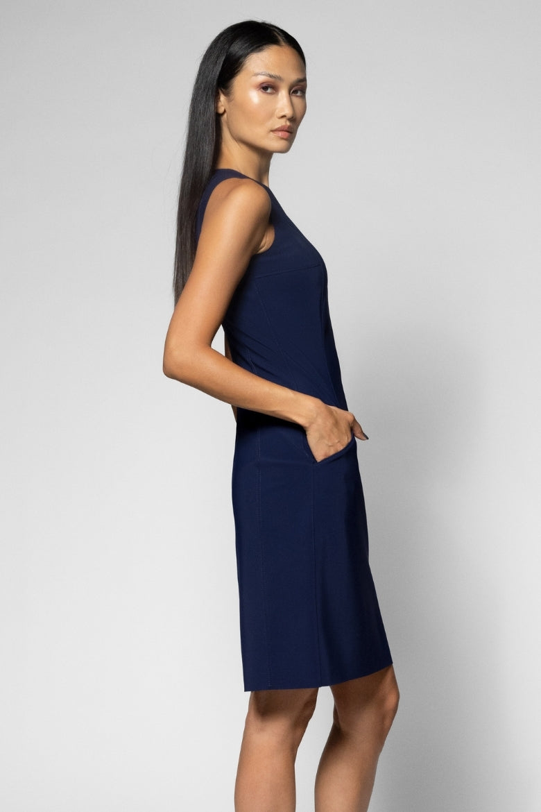 A woman modeling a sleeveless navy blue shift dress with side pockets, photographed against a light gray studio background.