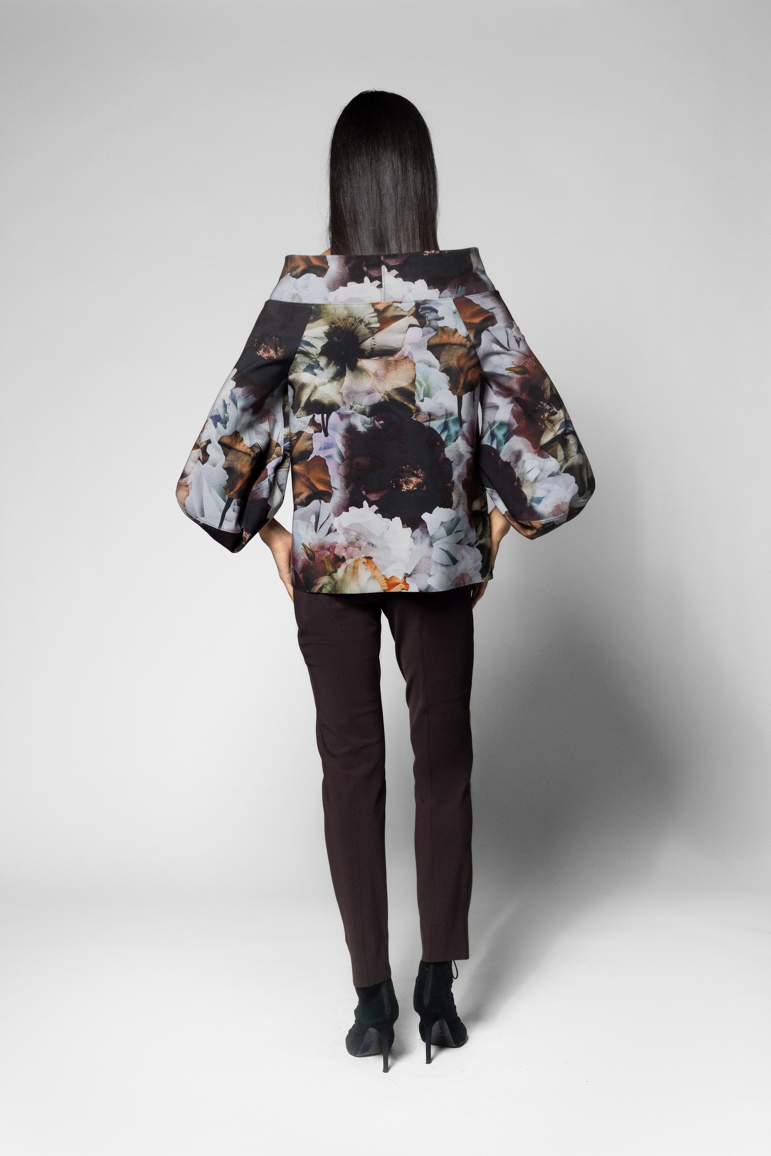 A model viewed from behind wearing a cropped floral print top with dramatic balloon sleeves, paired with dark brown trousers and black heels, against a light gray studio background.