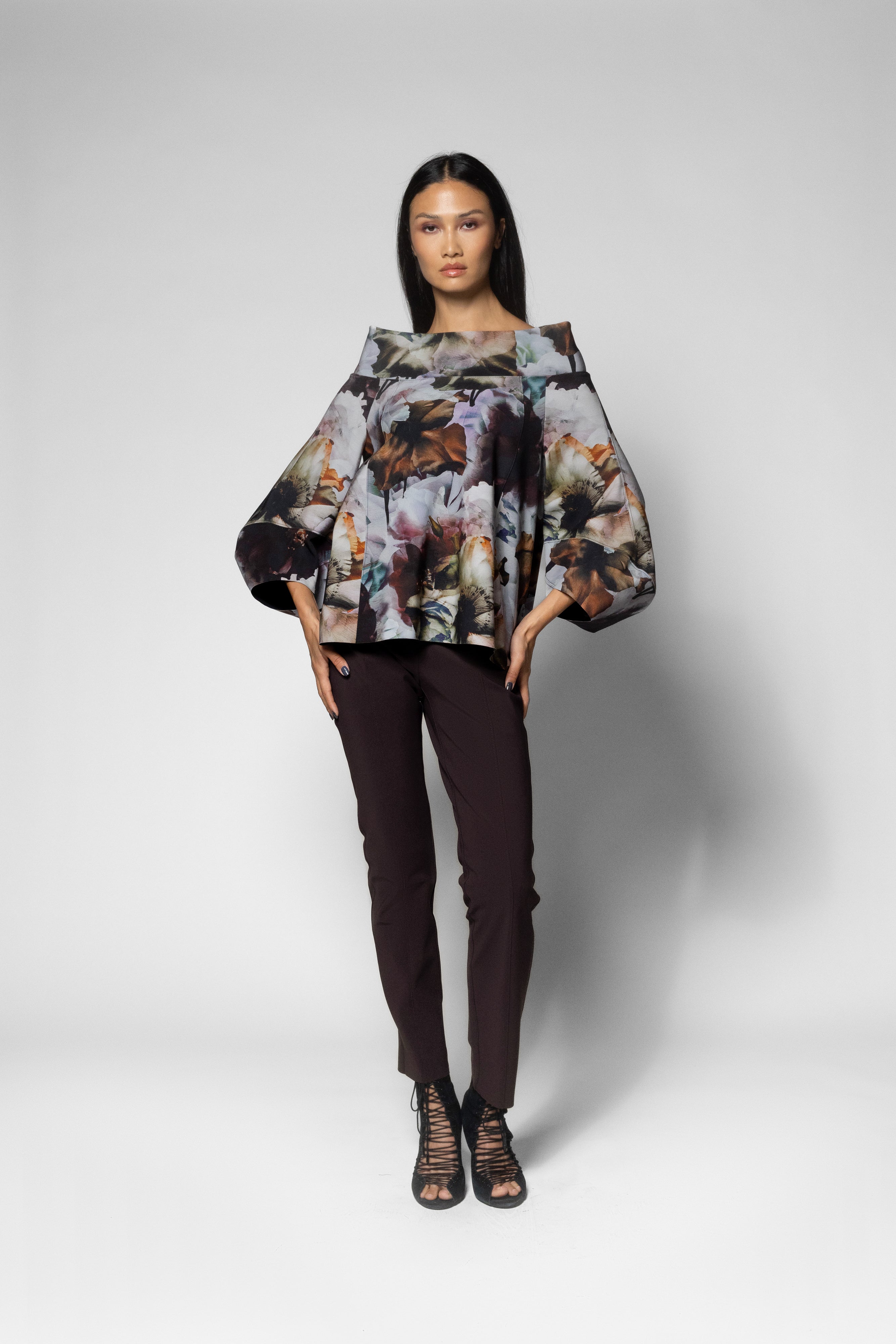 A model stands against a light gray studio background wearing a loose, oversized floral-patterned top with dramatic long sleeves, dark brown slim pants, and strappy high-heeled sandals.