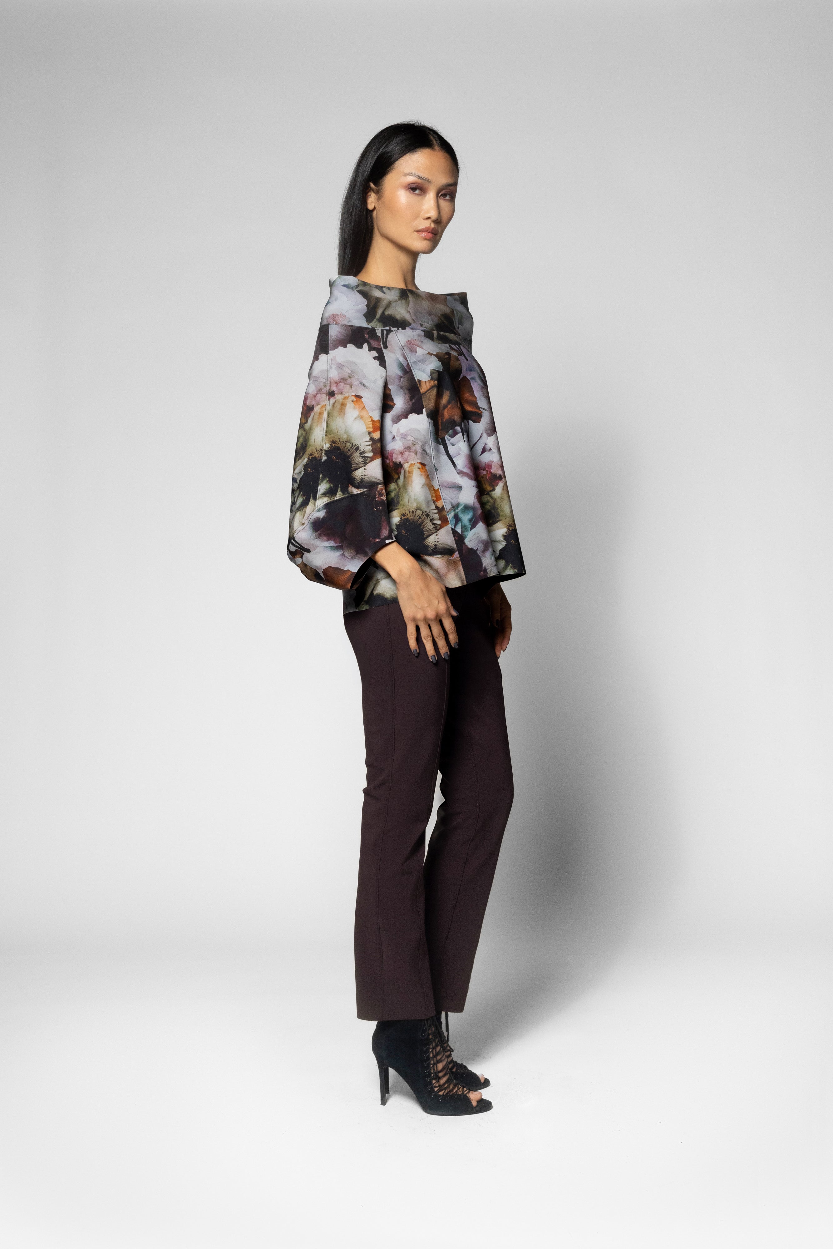 A model stands in profile wearing a dark floral print blouse with wide draped sleeves, dark brown trousers, and black lace-up high heels against a light gray studio background.