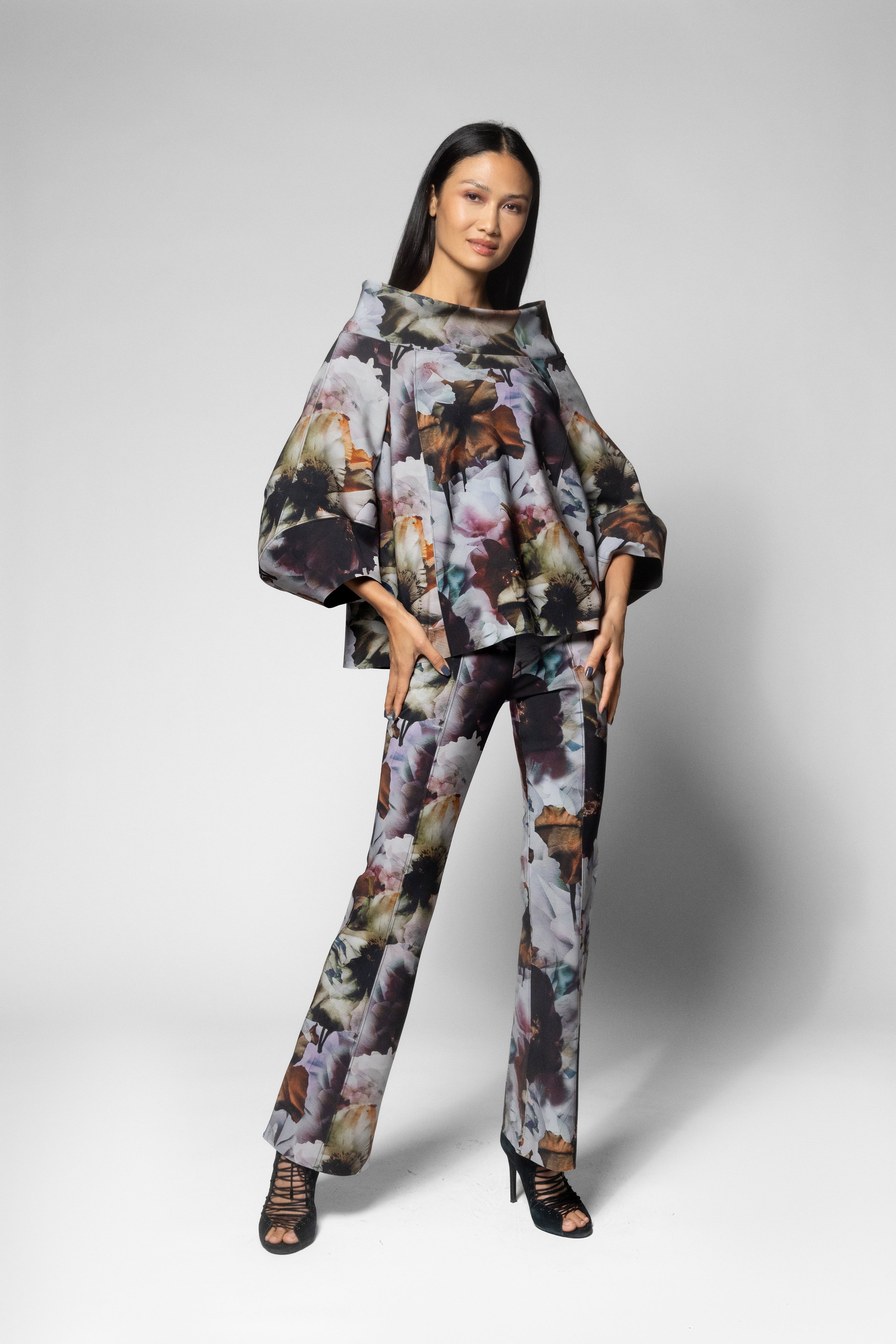 A model wearing a matching two-piece floral print set consisting of a wide-sleeved top and matching trousers, styled with black strappy high-heeled sandals against a light gray studio background.