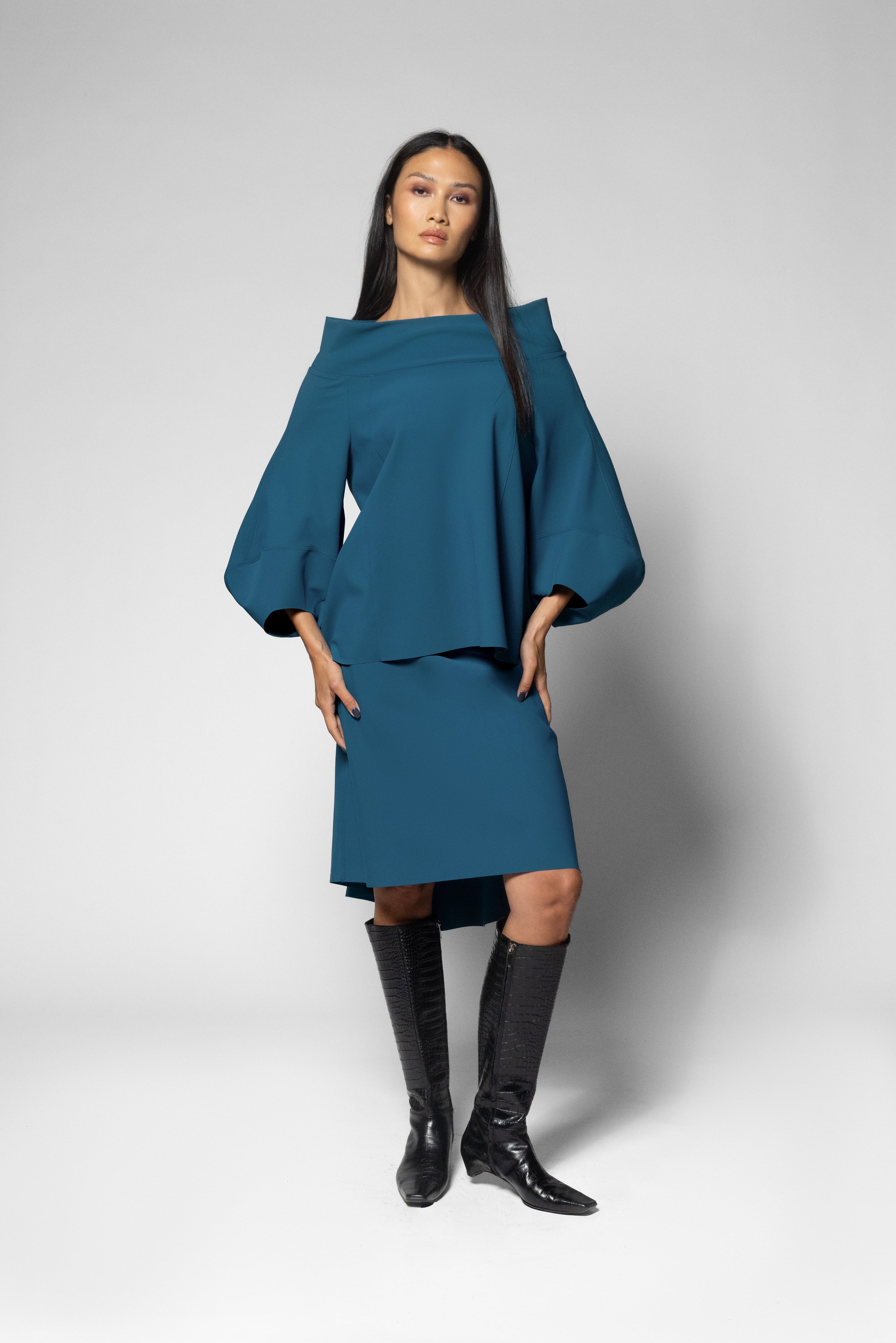 A model wearing a teal blue off-the-shoulder top with voluminous sleeves and a matching skirt, paired with black knee-high boots, against a light gray studio background.