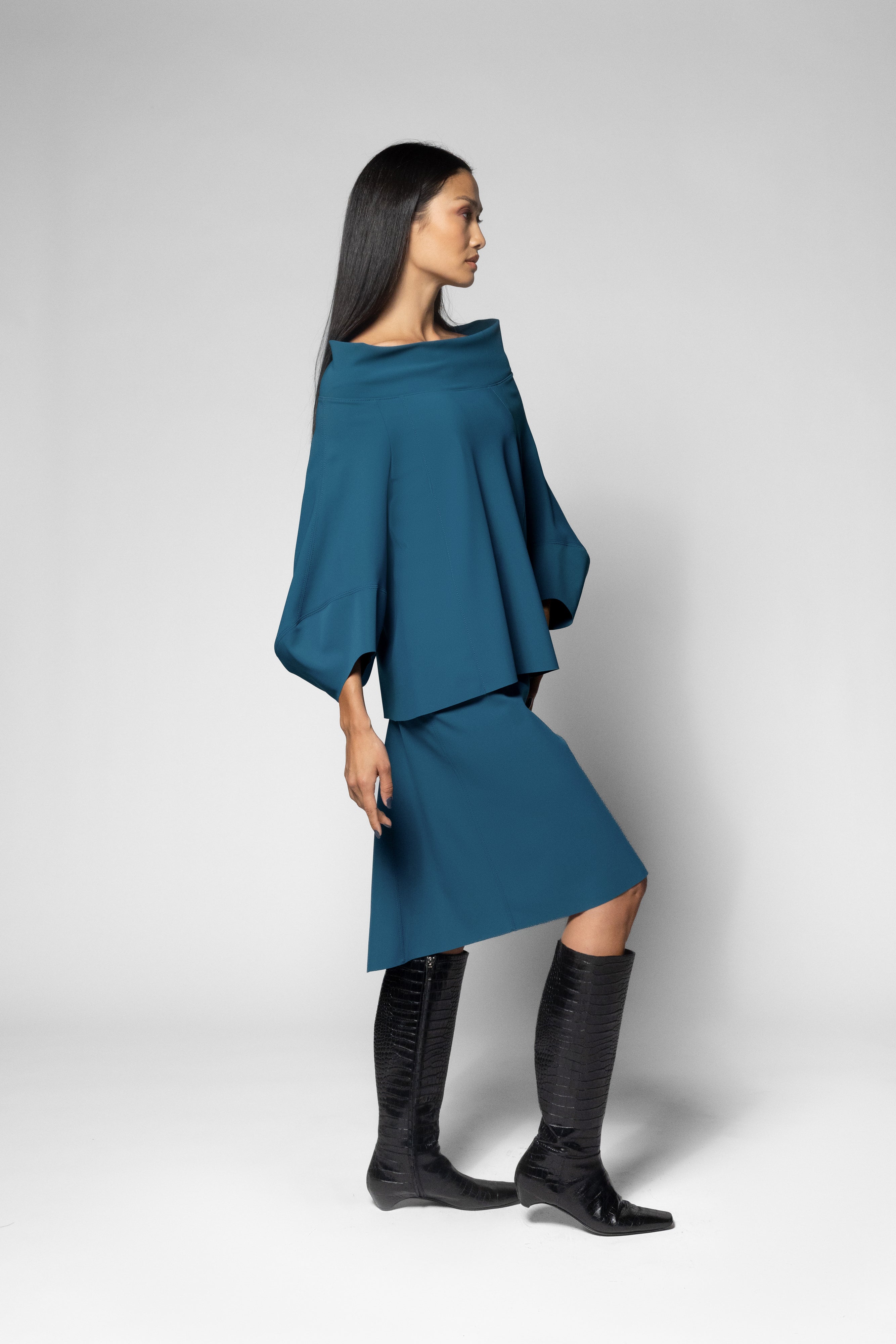 A woman modeling a teal blue off-the-shoulder top and a matching skirt with draped design and high-low hem, paired with black knee-high boots, against a light gray studio background.