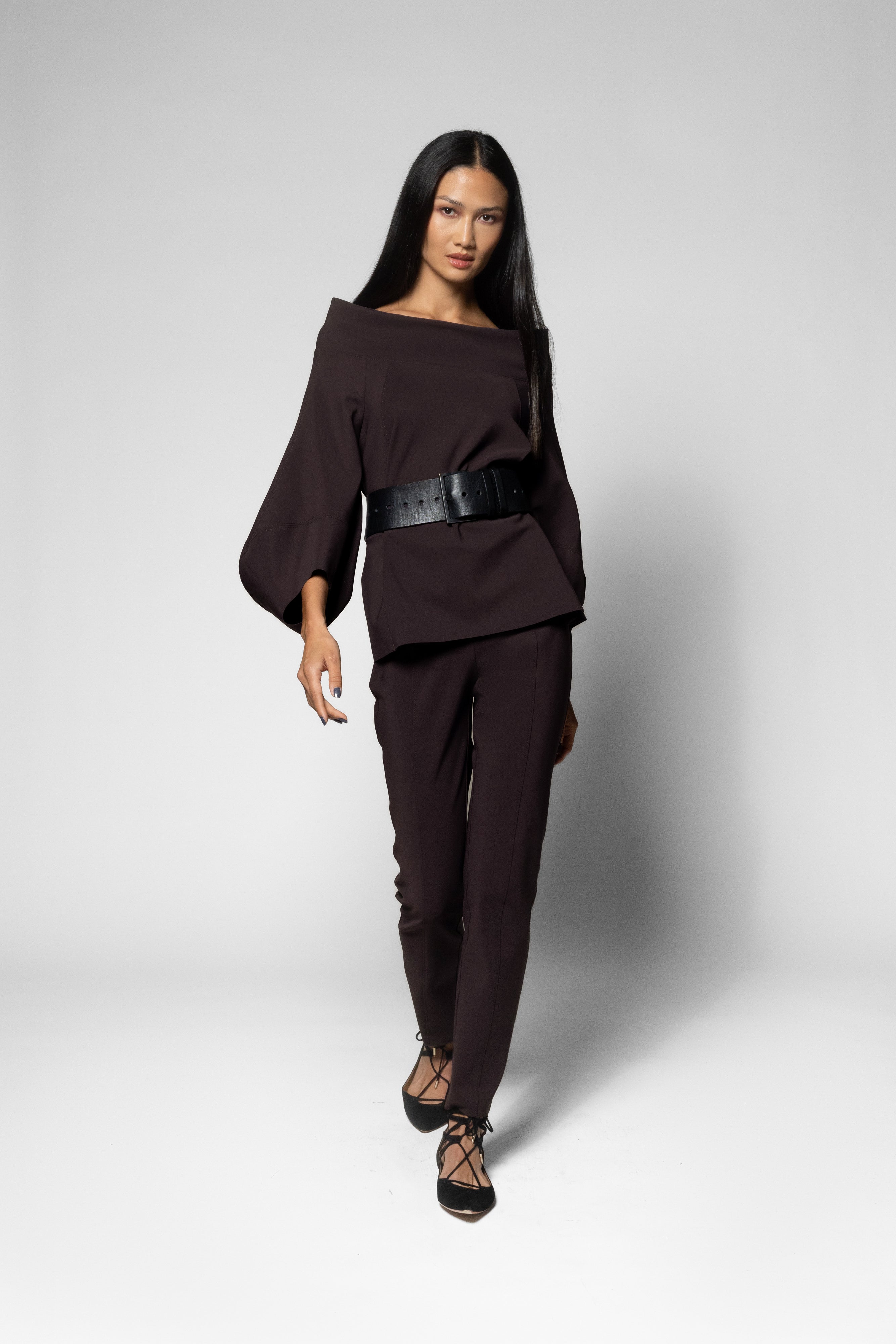 A model wearing a dark brown off-the-shoulder top with a wide black belt and matching dark brown trousers, walking against a light gray studio background.