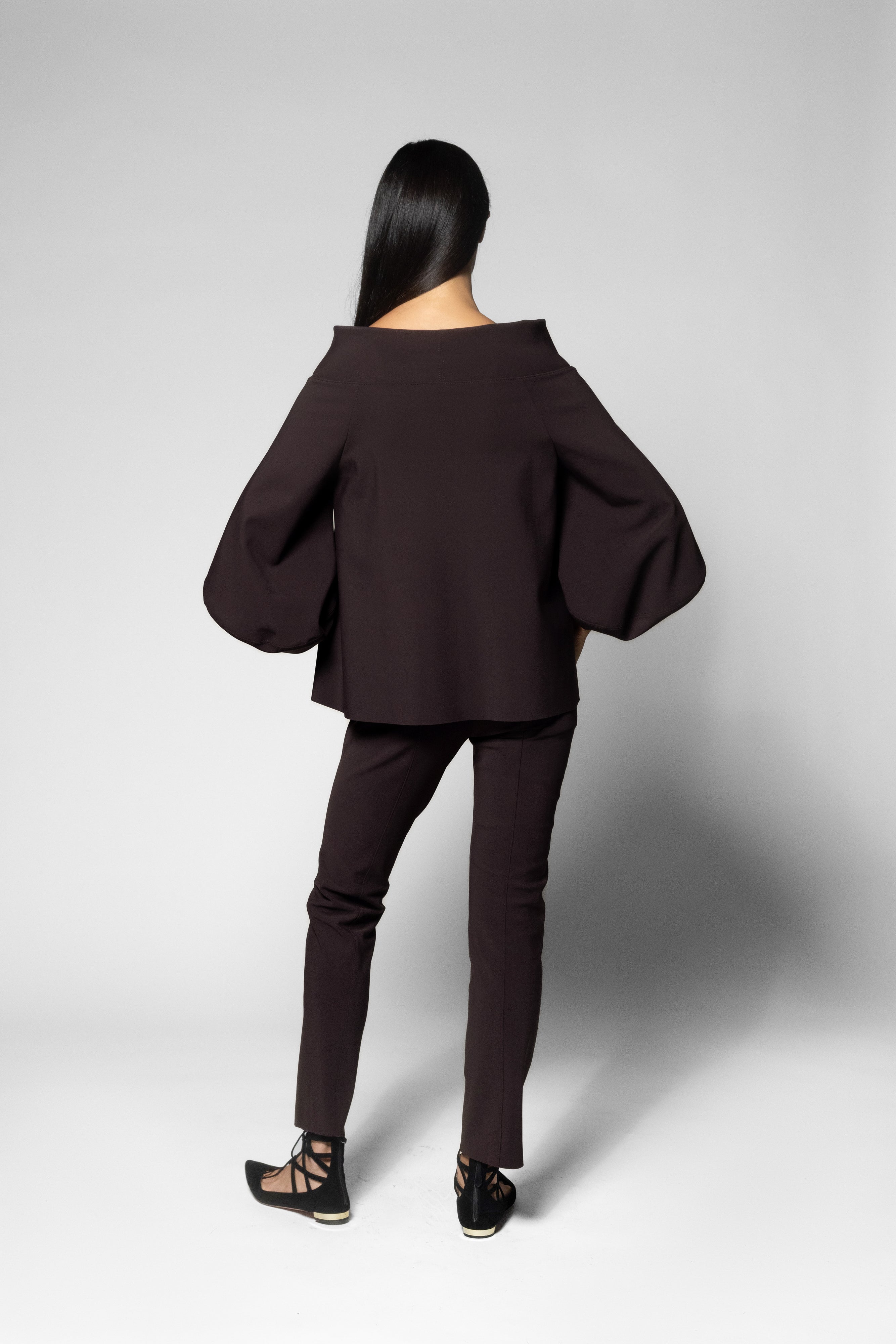 A model shown from the back wearing a dark brown off-the-shoulder top with dramatic balloon sleeves and matching dark brown slim-fit pants, set against a light gray studio background.