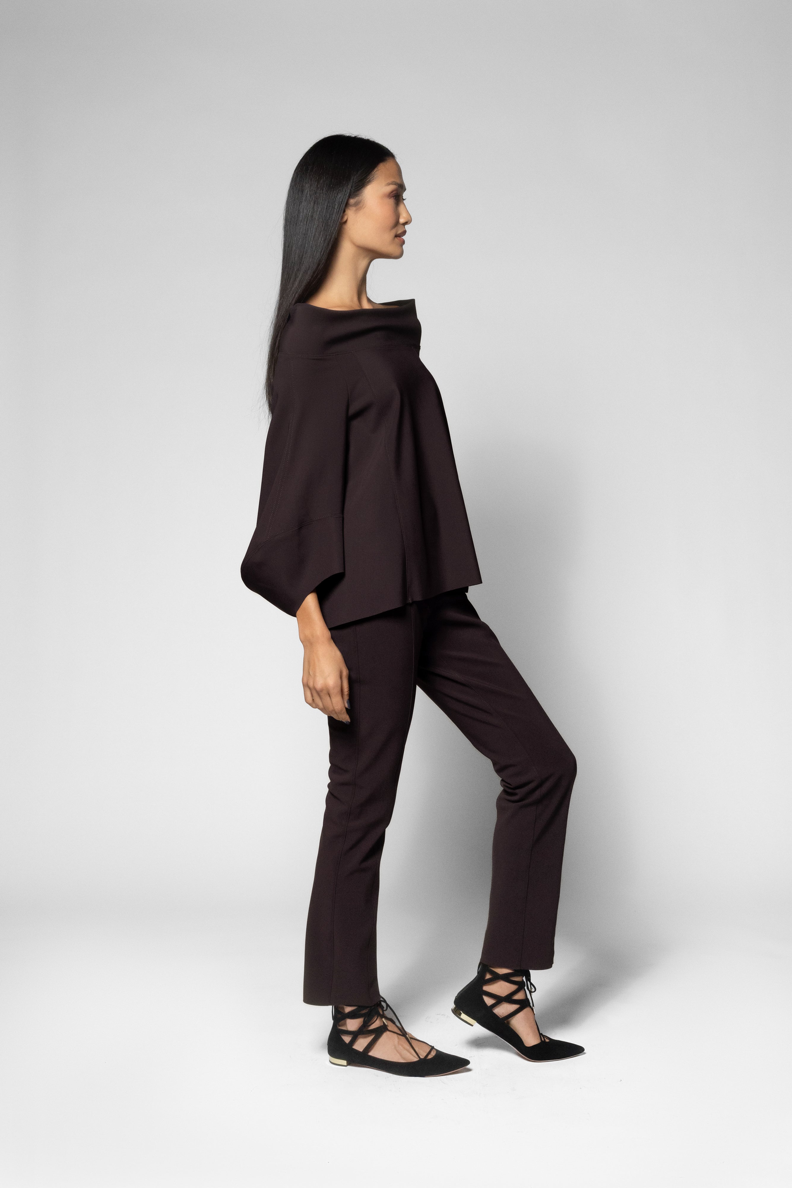 A woman modeling a dark brown two-piece outfit consisting of a draped, wide-sleeved top and matching straight-leg pants, paired with black strappy flat sandals against a light gray studio background.