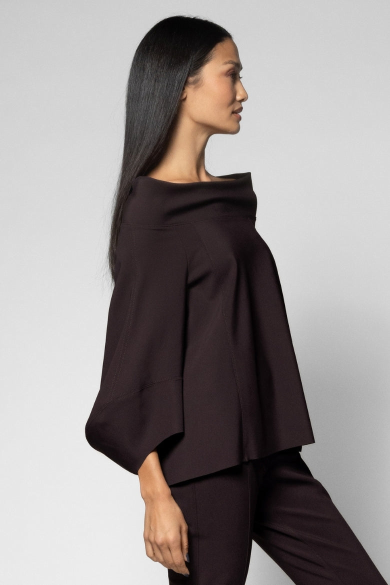 A woman in side profile wearing a dark brown off-the-shoulder draped top and matching dark brown trousers, photographed against a light gray studio background.