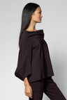 A woman in side profile wearing a dark brown off-the-shoulder draped top and matching dark brown trousers, photographed against a light gray studio background.