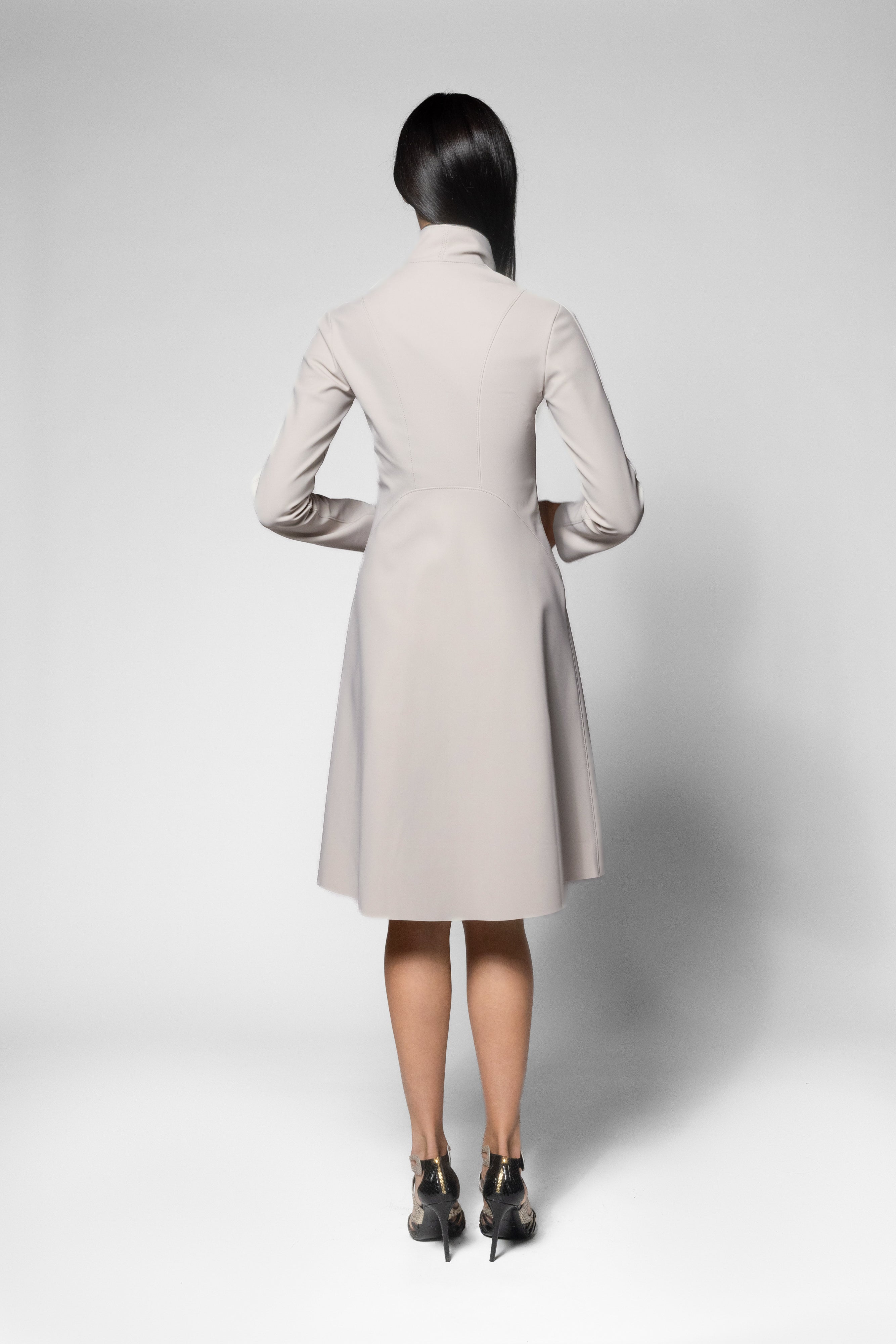 A woman viewed from the back wearing a light beige long-sleeve knee-length coat , standing against a plain light gray studio background.
