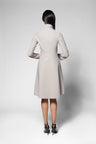 A woman viewed from the back wearing a light beige long-sleeve knee-length coat , standing against a plain light gray studio background.