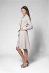 A woman modeling a light beige long-sleeve wrap-style coat dress against a plain light gray studio background.