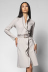 A woman modeling a light gray wrap-style coat with a wide collar, long sleeves, and a textured python belt, photographed against a plain light gray studio background.