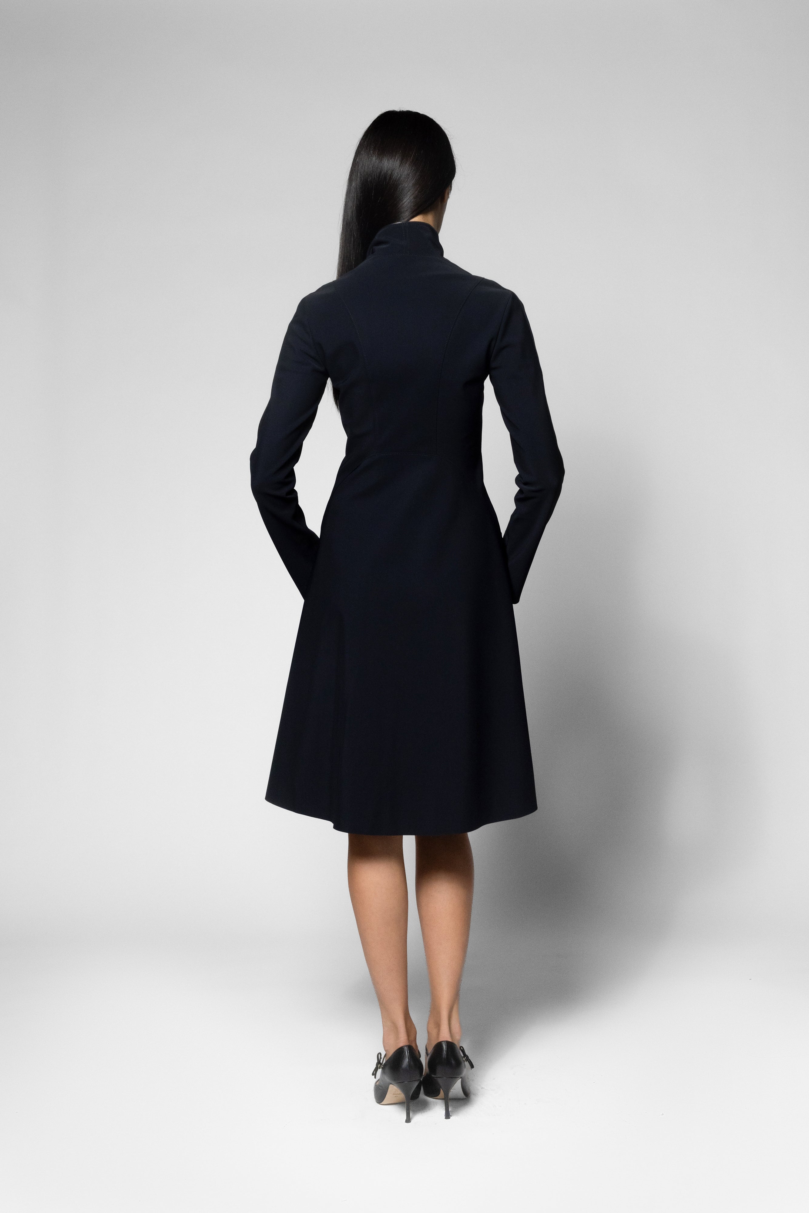 A woman viewed from the back wearing a black long-sleeve knee-length coat with a high collar and hands in pockets, standing against a light gray studio background.