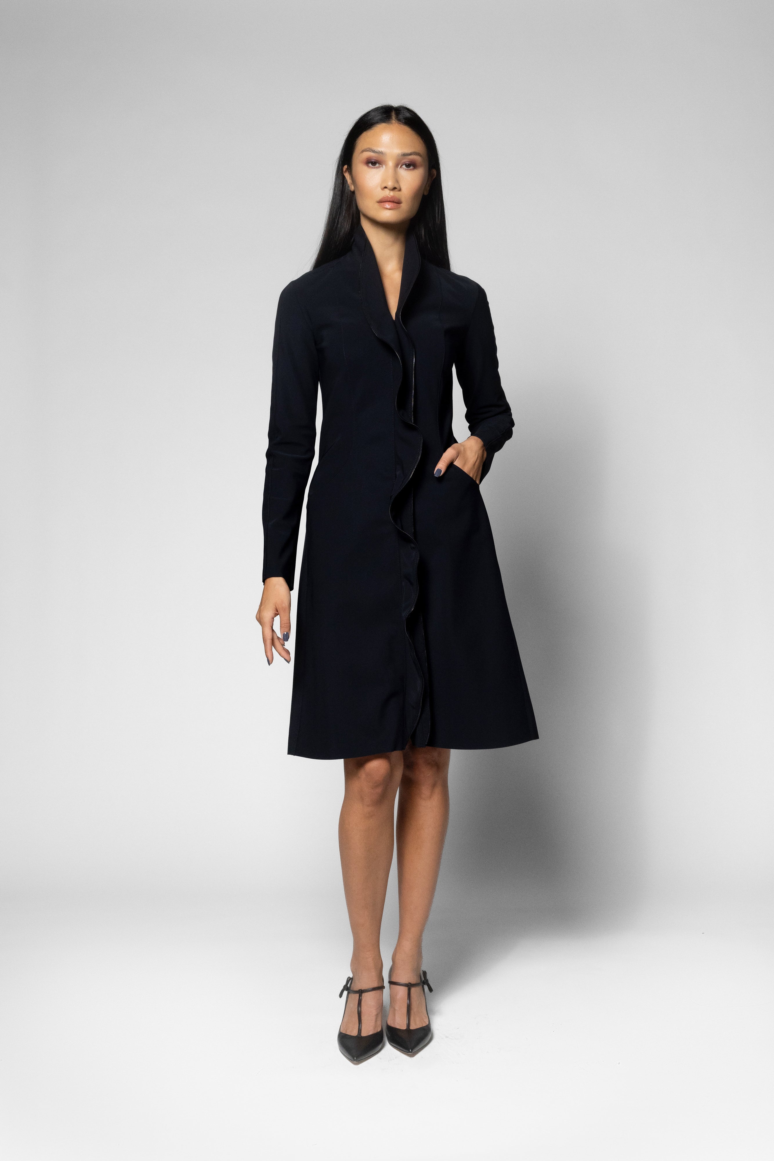 A woman modeling a black long-sleeve  coat with a V-neckline and front pockets, standing against a light gray studio background.