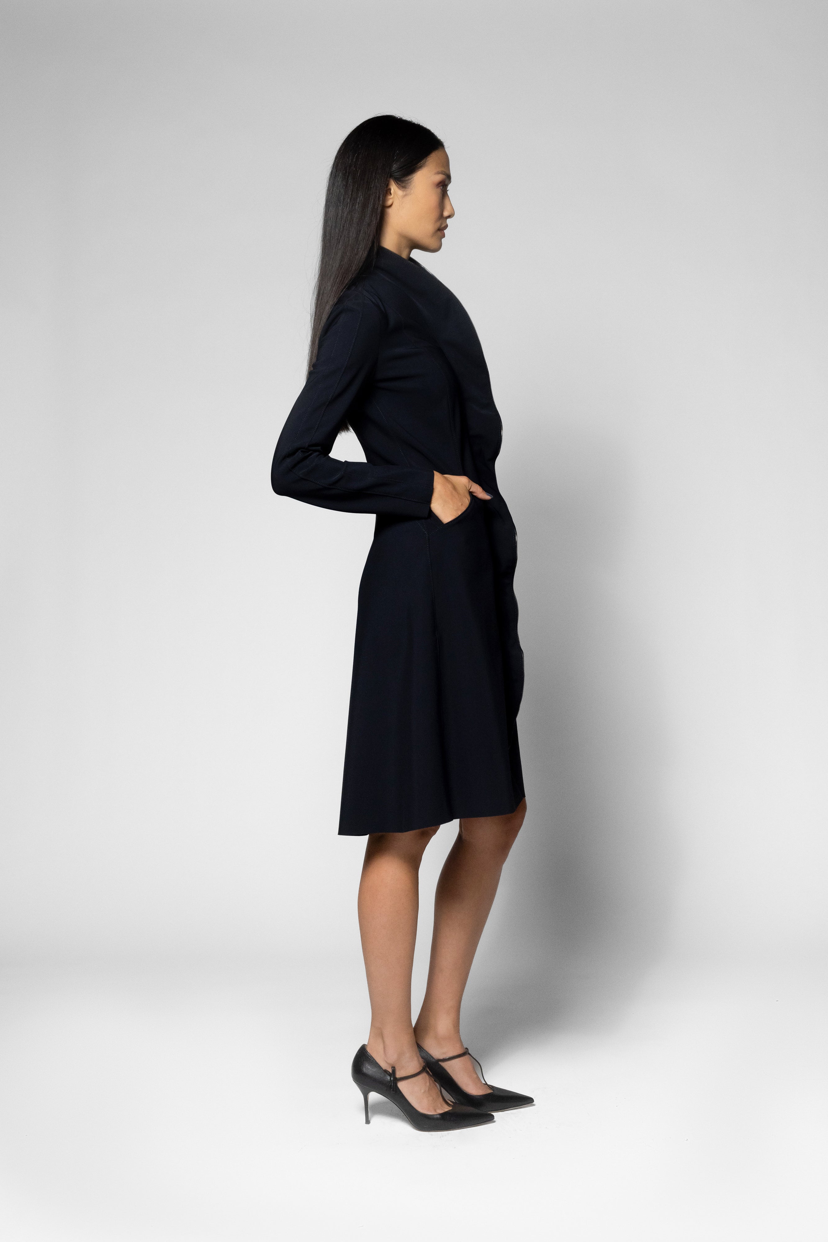A woman modeling a black long-sleeve knee-length coat, shown from a side profile against a light gray studio background.
