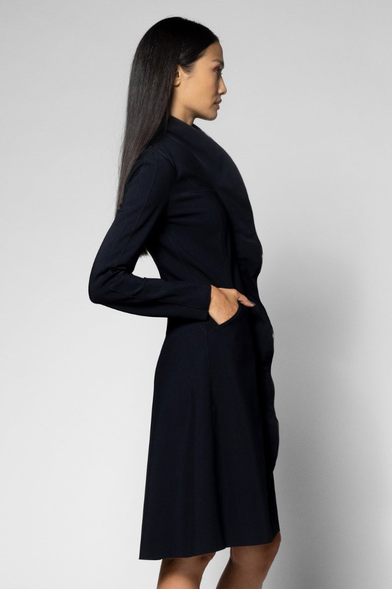 A woman in profile wearing a black long-sleeve coat against a light gray studio background.