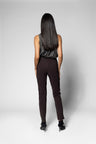 A woman viewed from behind wearing slim dark brown pants, a textured dark gray sleeveless top, and black pointed-toe heels against a light gray studio background.