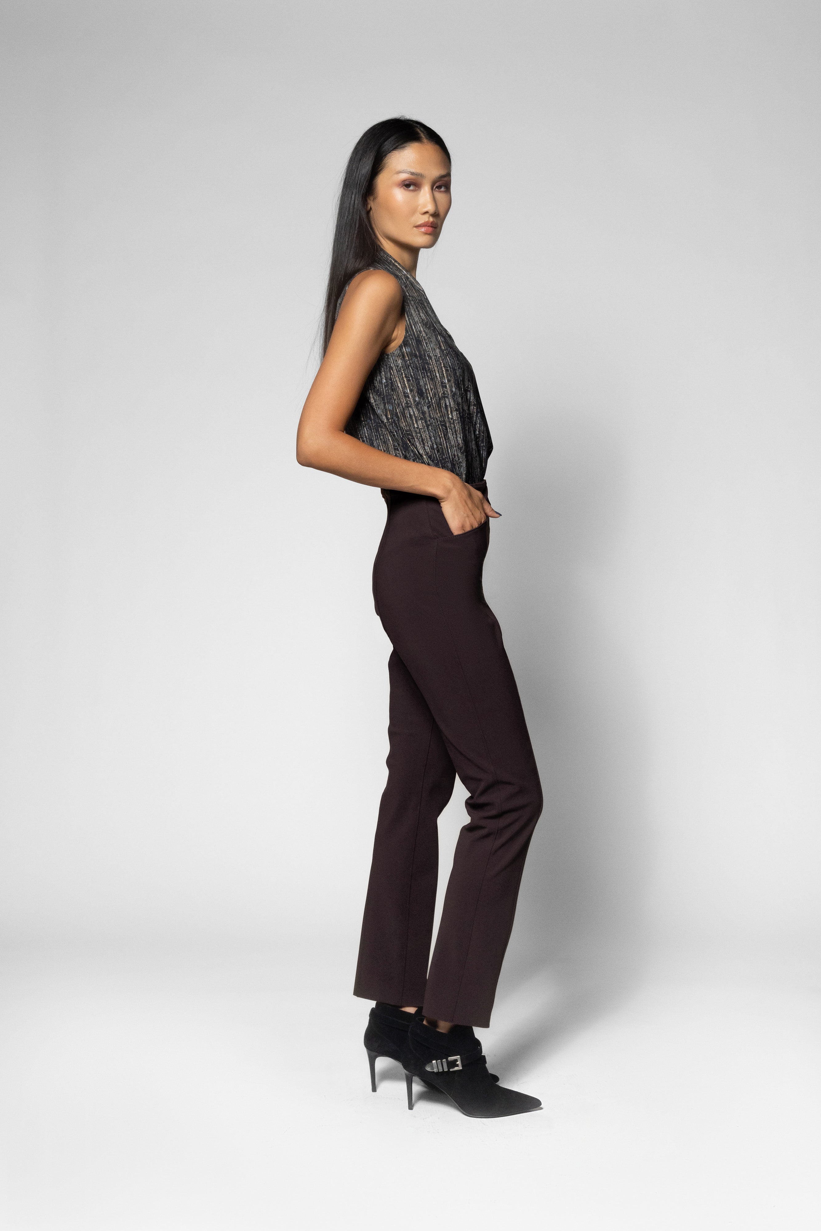 A model is shown from the side in a studio setting, wearing a sleeveless dark gray patterned top, dark brown trousers, and black high-heeled shoes against a light gray background.