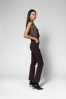 A model is shown from the side in a studio setting, wearing a sleeveless dark gray patterned top, dark brown trousers, and black high-heeled shoes against a light gray background.