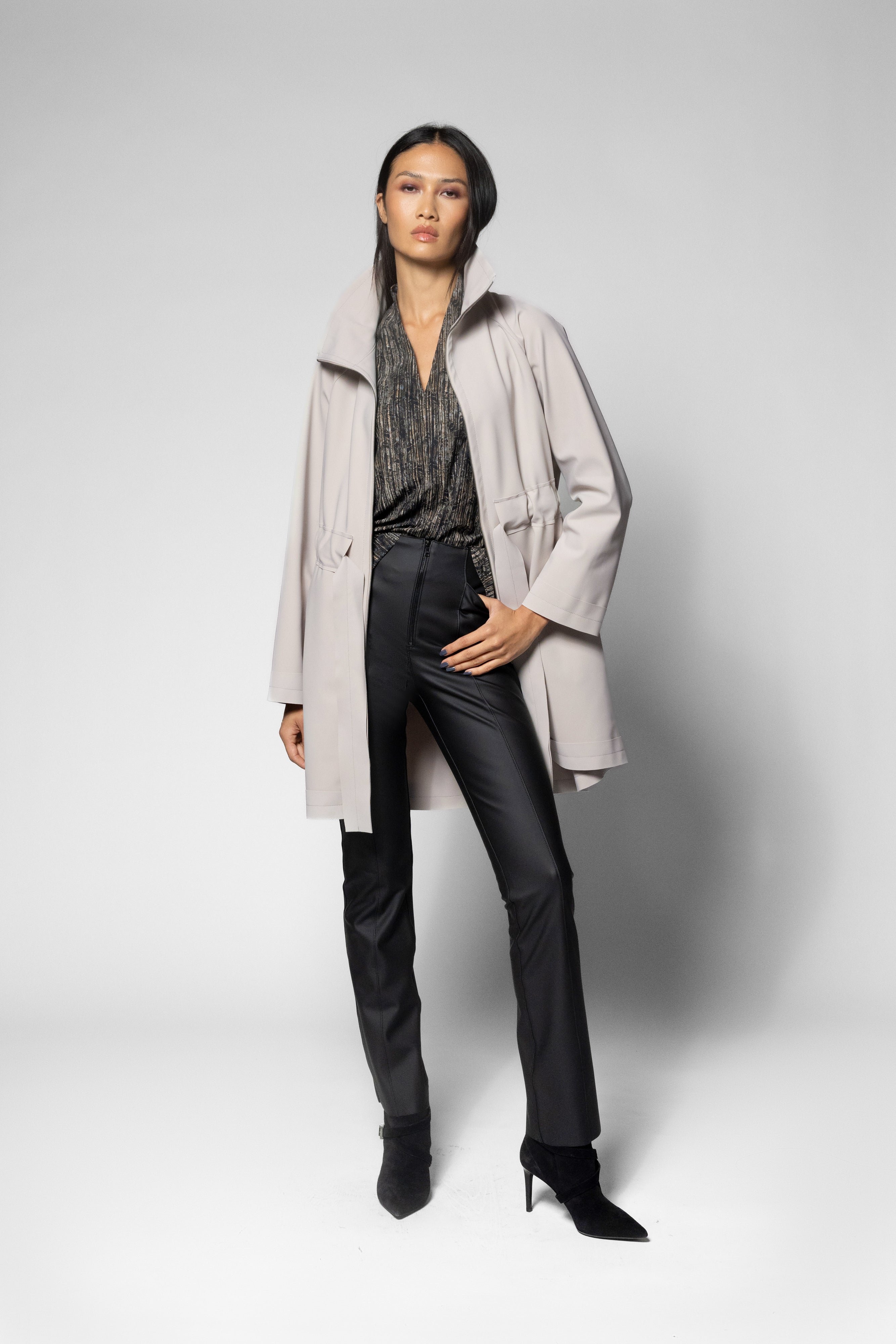 A model stands in a studio wearing a light beige trench coat over a dark patterned blouse, black trousers, and black pointed-toe heels against a light gray background.