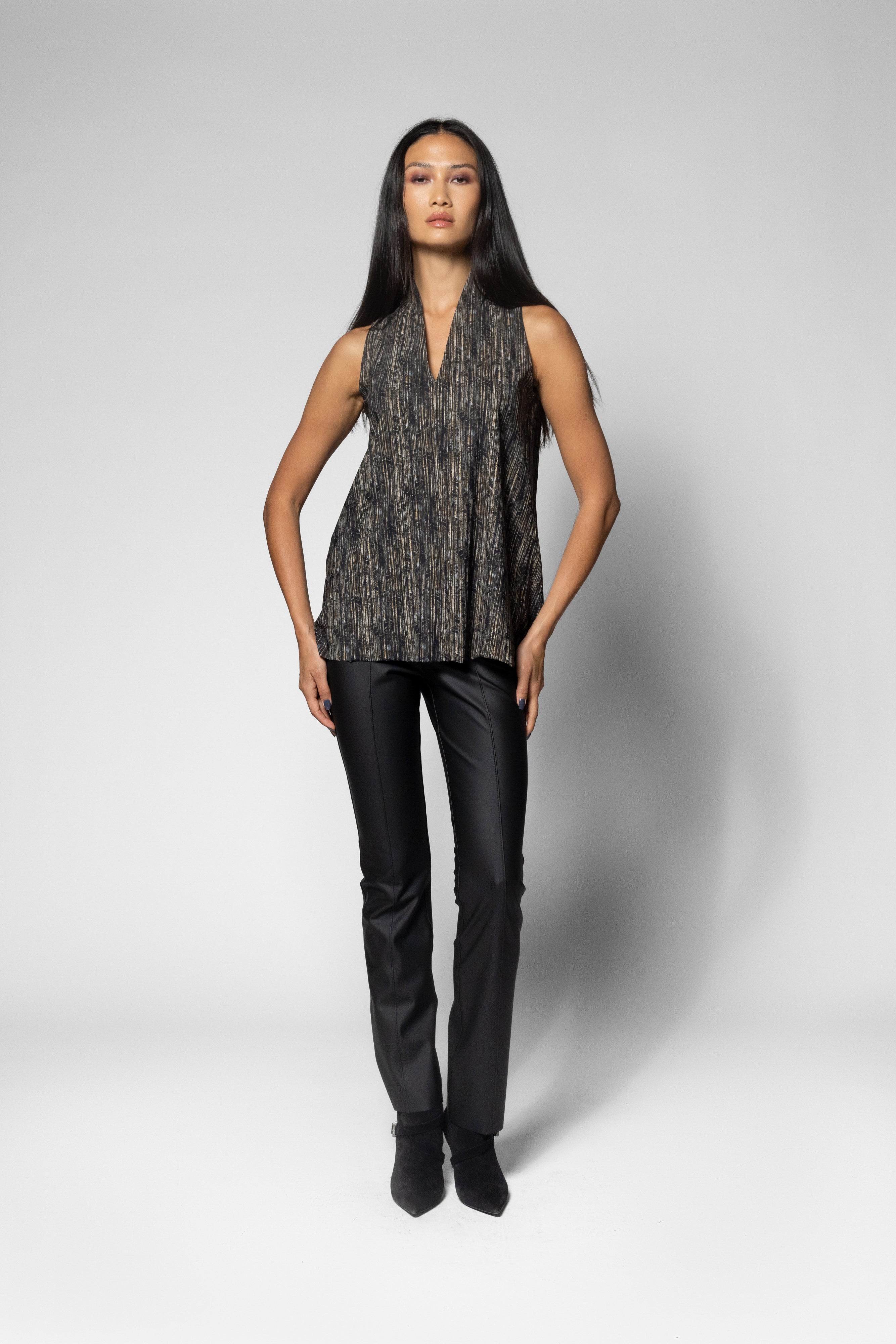 A model stands against a light gray studio background wearing a sleeveless textured top with a dark gray and black mottled pattern, paired with black pants and black ankle boots.