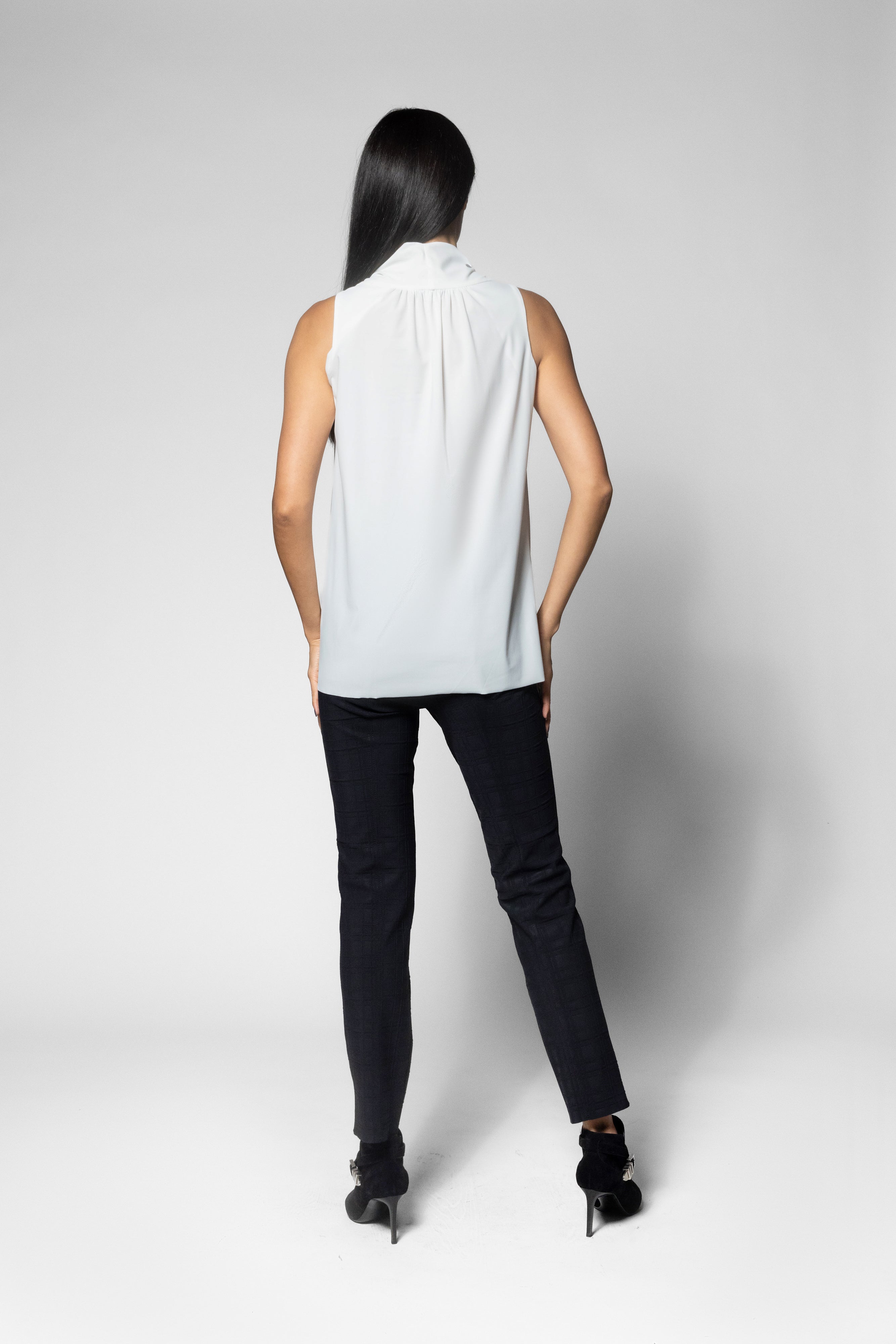 A woman shown from the back wearing a sleeveless white blouse with a gathered high neck, black pants, and black heels, photographed against a light gray studio background.