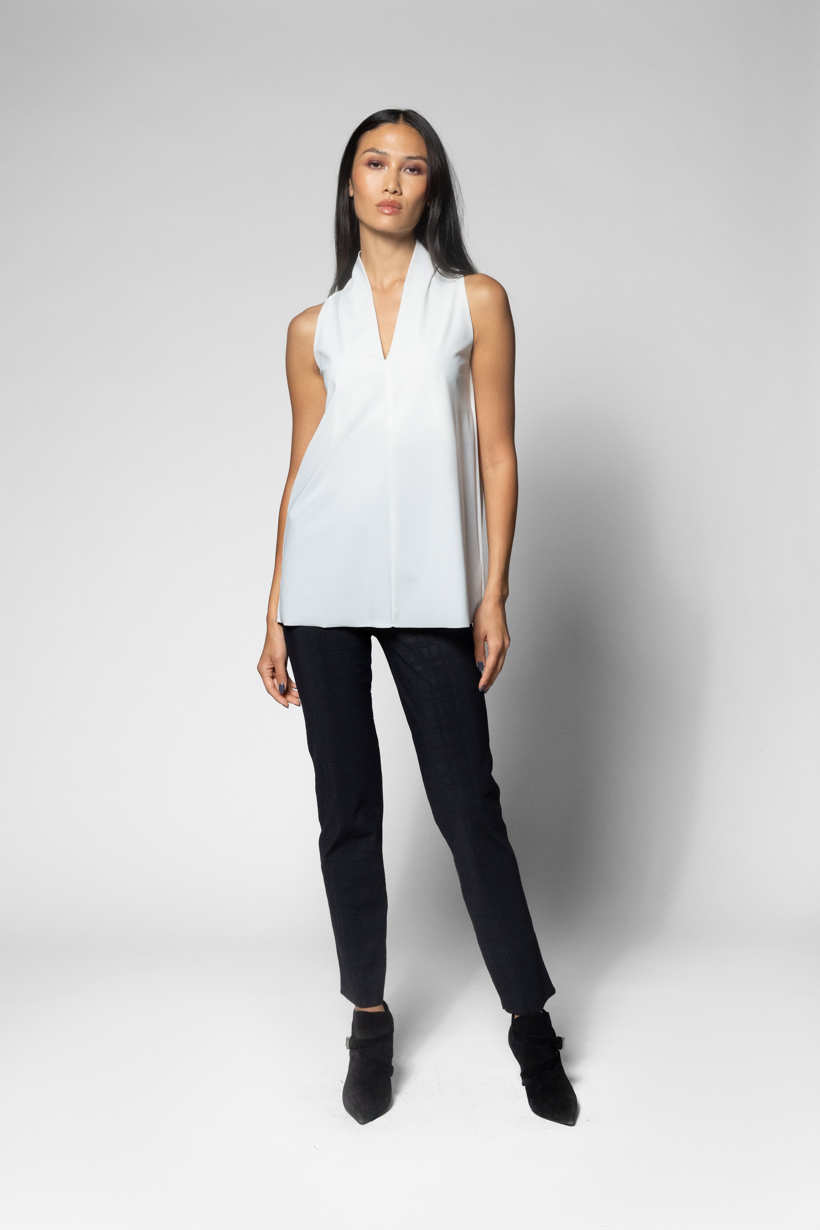 A model stands against a light gray studio background wearing a sleeveless white wrap-style top, black pants, and black ankle boots.