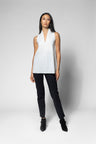 A model stands against a light gray studio background wearing a sleeveless white wrap-style top, black pants, and black ankle boots.