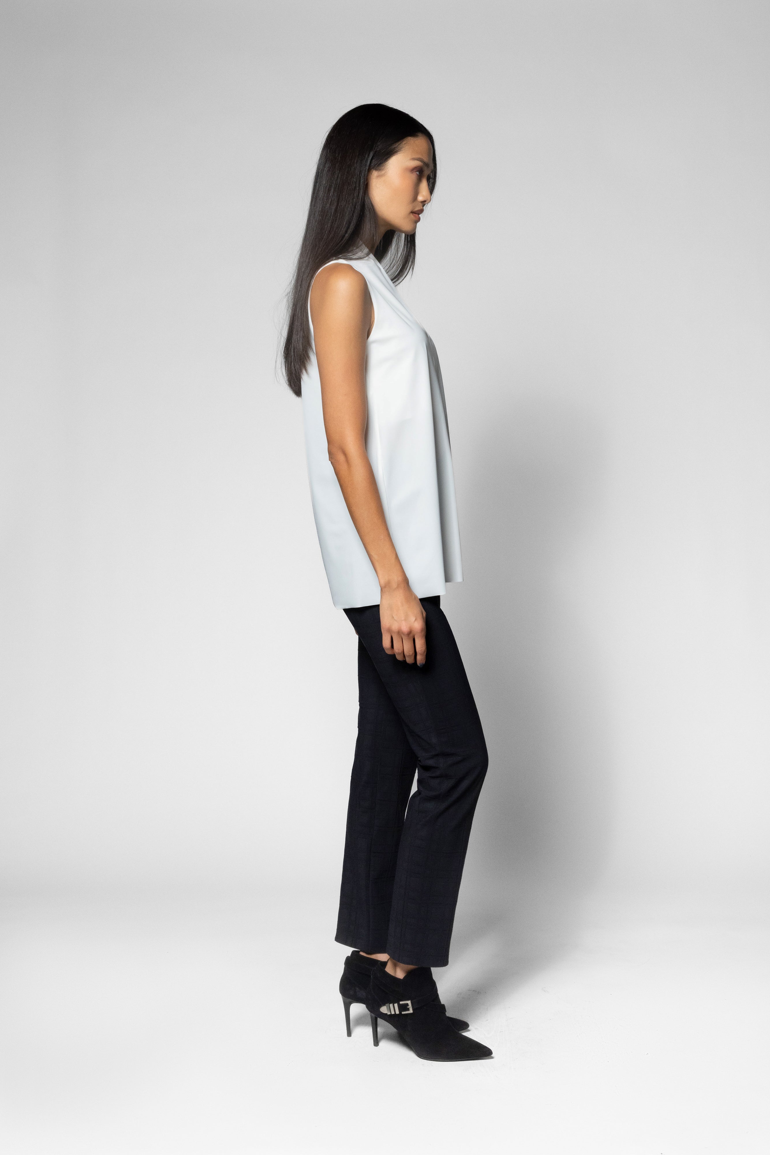 A woman in side profile wearing a sleeveless white top, black wide-leg trousers, and black high-heeled shoes against a light gray studio background.