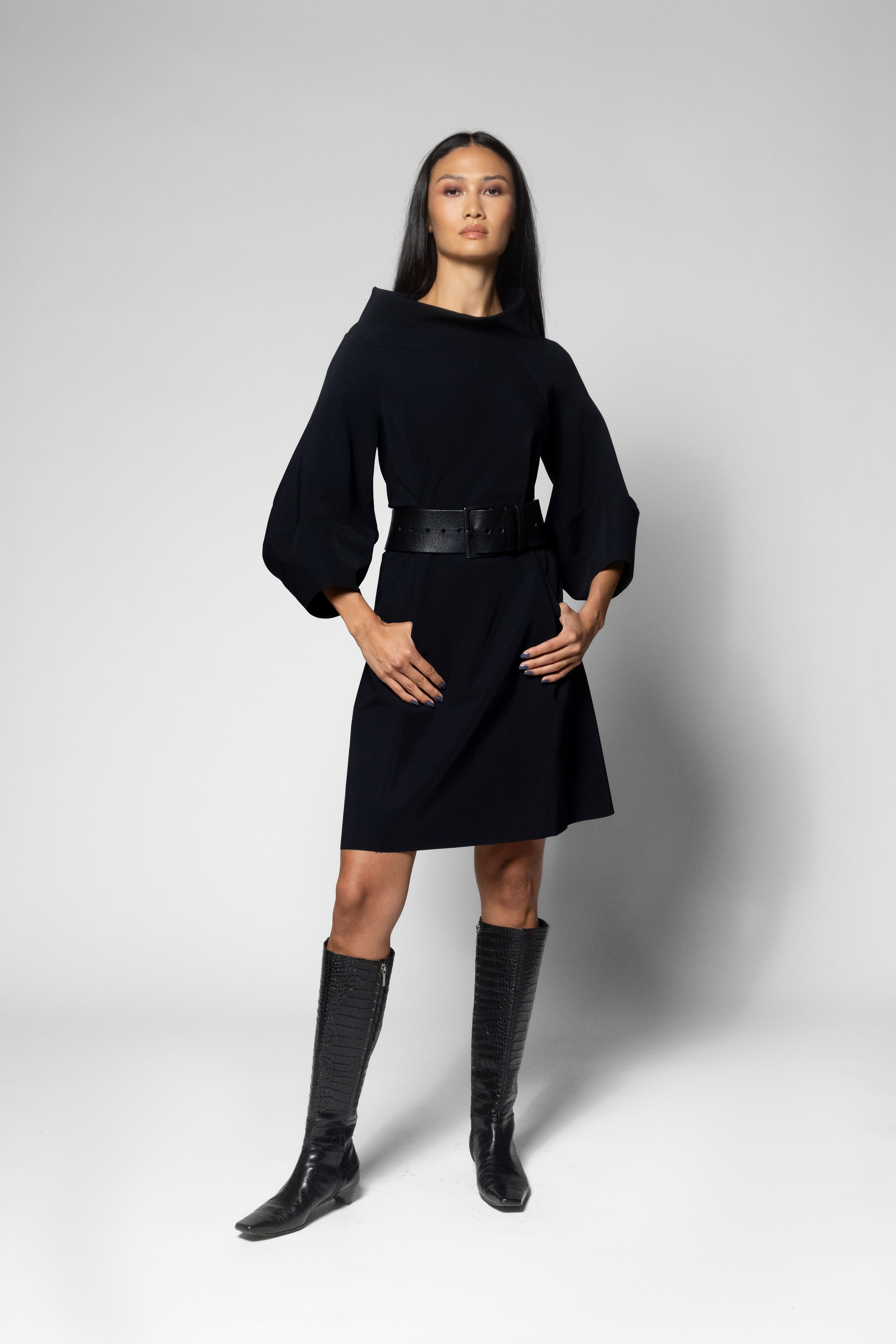 A woman modeling a black long-sleeve mini dress with a wide black belt, paired with black knee-high boots, against a light gray studio background.
